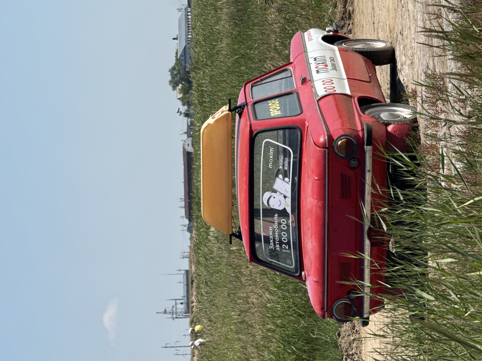 A red and white vintage car, likely a taxi given the branding, is parked on a dirt road surrounded by tall green grass. The car has a yellow rack on its roof. On the rear window, there is a sticker that reads "Закажи автомобиль 2 00 00" which translates to "Order a car 2 00 00". Another sticker on the window says "maxim". The car also has "00 00 maxim" printed on its side with the Russian word "Заказки такси" which means "Taxi orders". In the background, there are power lines, some buildings, and two people walking in a field. The sky is clear and blue. The scene appears to be in a rural or semi-rural area, possibly on the outskirts of Bursool, Russia.