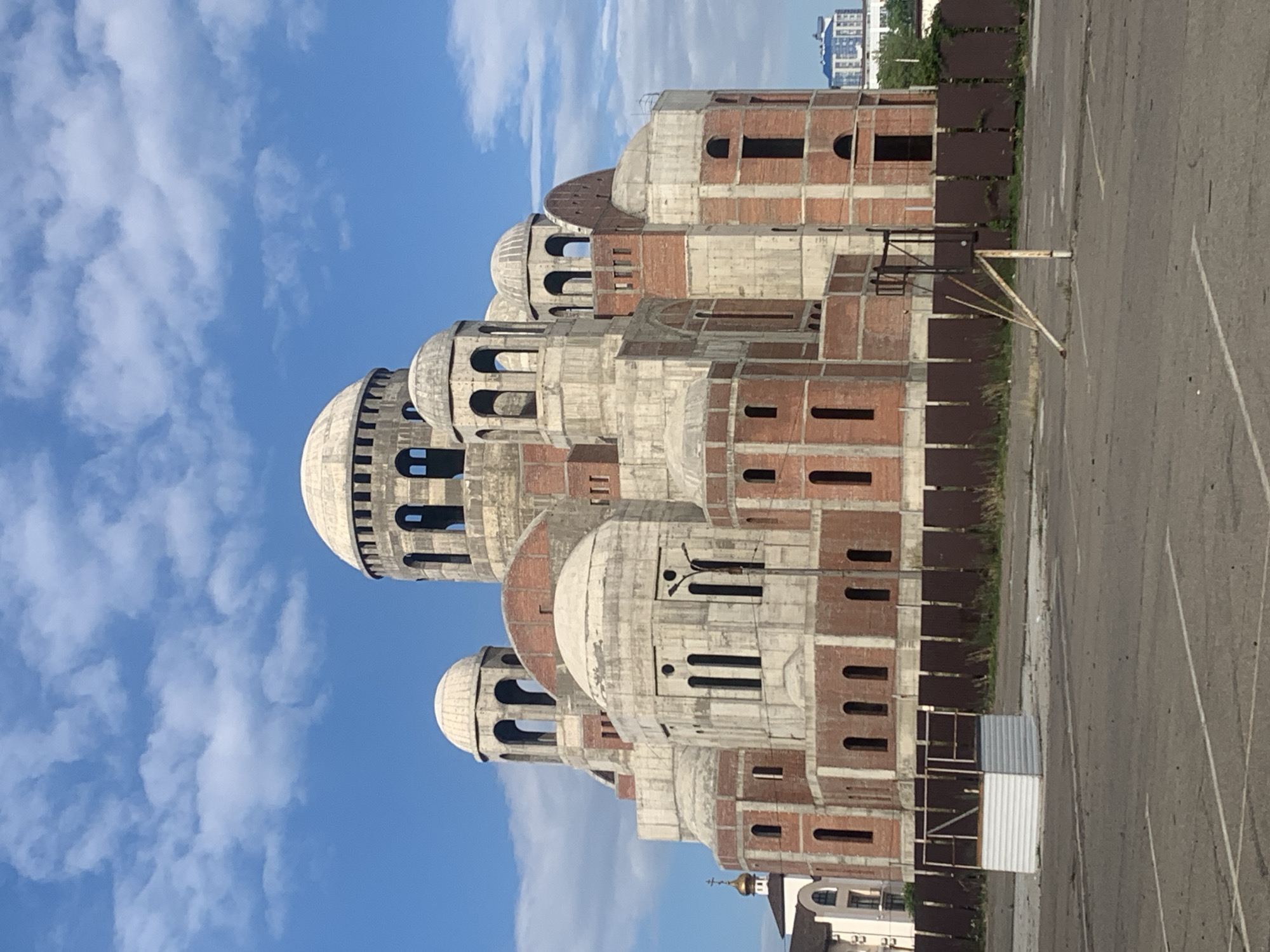 The image shows the unfinished construction of a large, domed building, likely a church, in Novorossiysk, Russia. The structure is primarily composed of exposed red brick and concrete, with multiple domes and arched windows in various stages of completion. Several smaller domes and bell towers are visible, as well as a central, larger dome. The building appears to be surrounded by a chain-link fence and a brown wooden fence. In the foreground, there is an asphalt surface with parking lines, and a dilapidated basketball hoop. A small white utility structure with a metal frame is also present in the foreground. The sky is blue with scattered white clouds. In the background, a smaller church with a golden dome is visible to the left, and a modern building with blue windows can be seen further in the distance to the right.