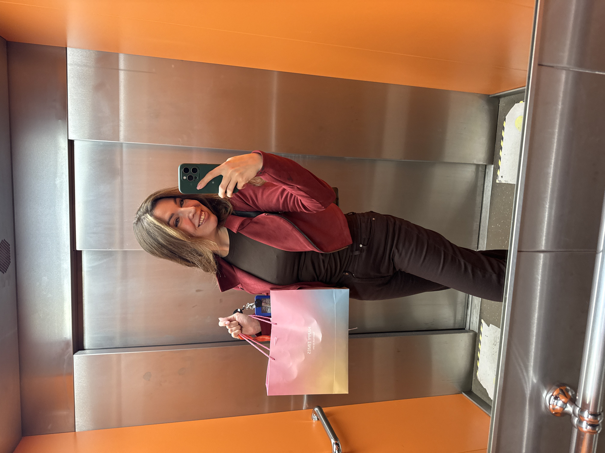 A young woman takes a selfie in what appears to be an elevator. She is holding a shopping bag and a smartphone. The elevator's interior is characterized by stainless steel and orange panels. The image does not provide sufficient details to determine if the location is Kryvyi Rih, Ukraine.