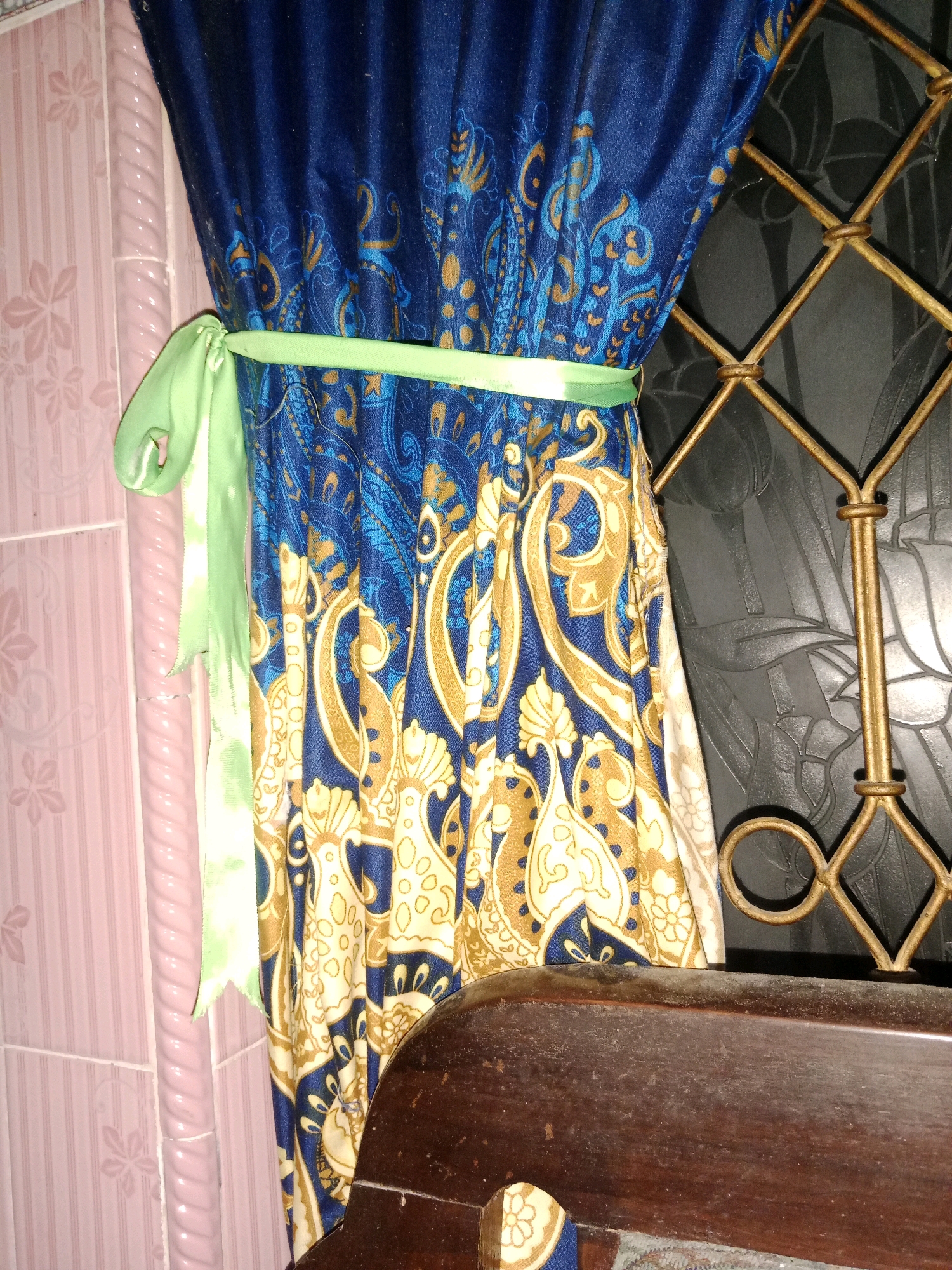 A close-up, eye-level shot showcases a blue and gold patterned curtain tied back with a light green sash against a textured pink wall and a dark, ornate metal grate, with the edge of a dark wooden object in the foreground. The scene is situated in Ringinrejo, Indonesia.