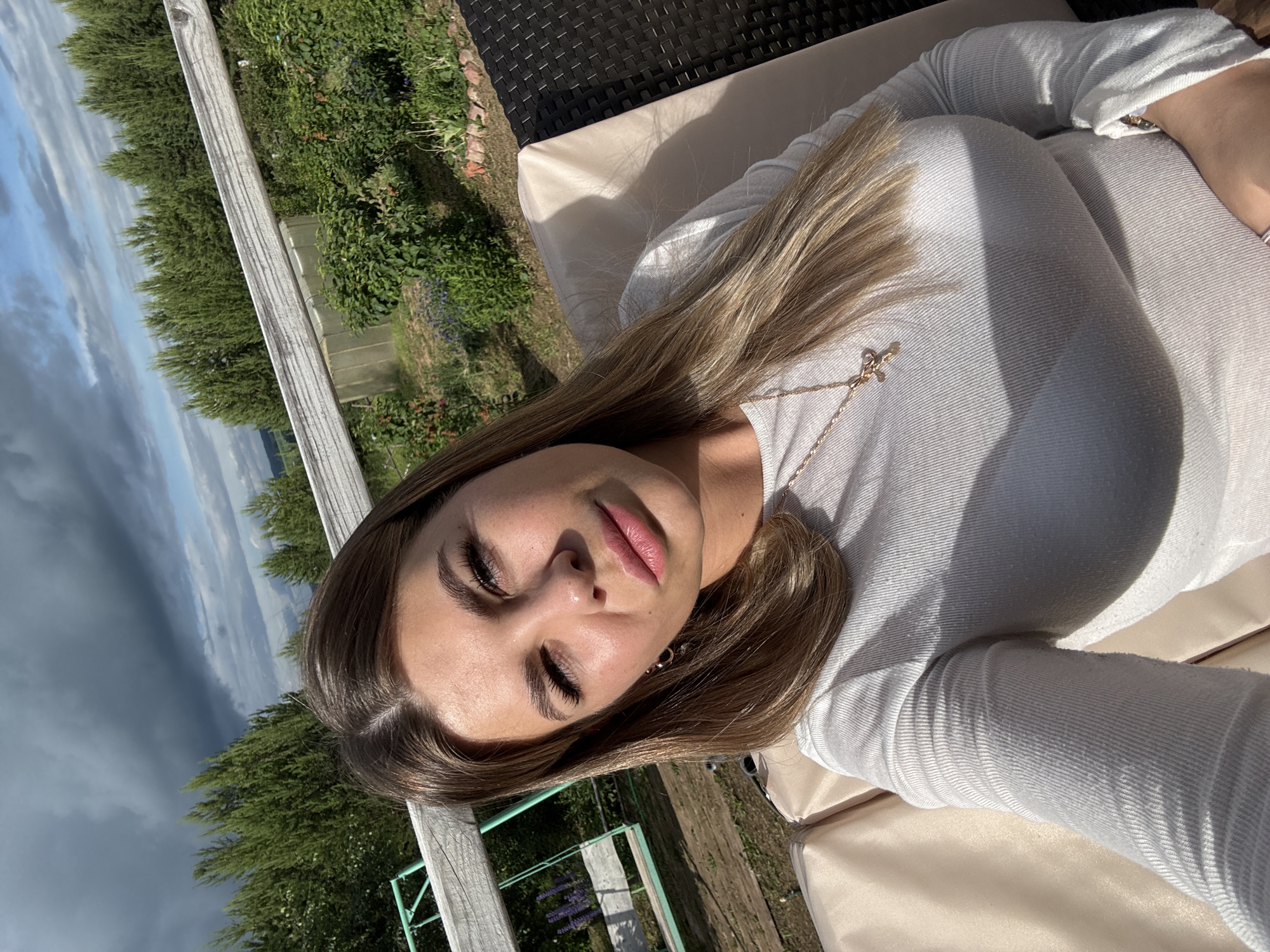 A woman with blonde hair, wearing a white long-sleeved shirt and a gold necklace, is taking a selfie outdoors. She is seated on a beige outdoor couch, with a dark rattan chair to her right. In the background, there is a wooden railing, green trees, and a cloudy sky. A swing set is visible to the left. The location appears to be a yard or garden area.