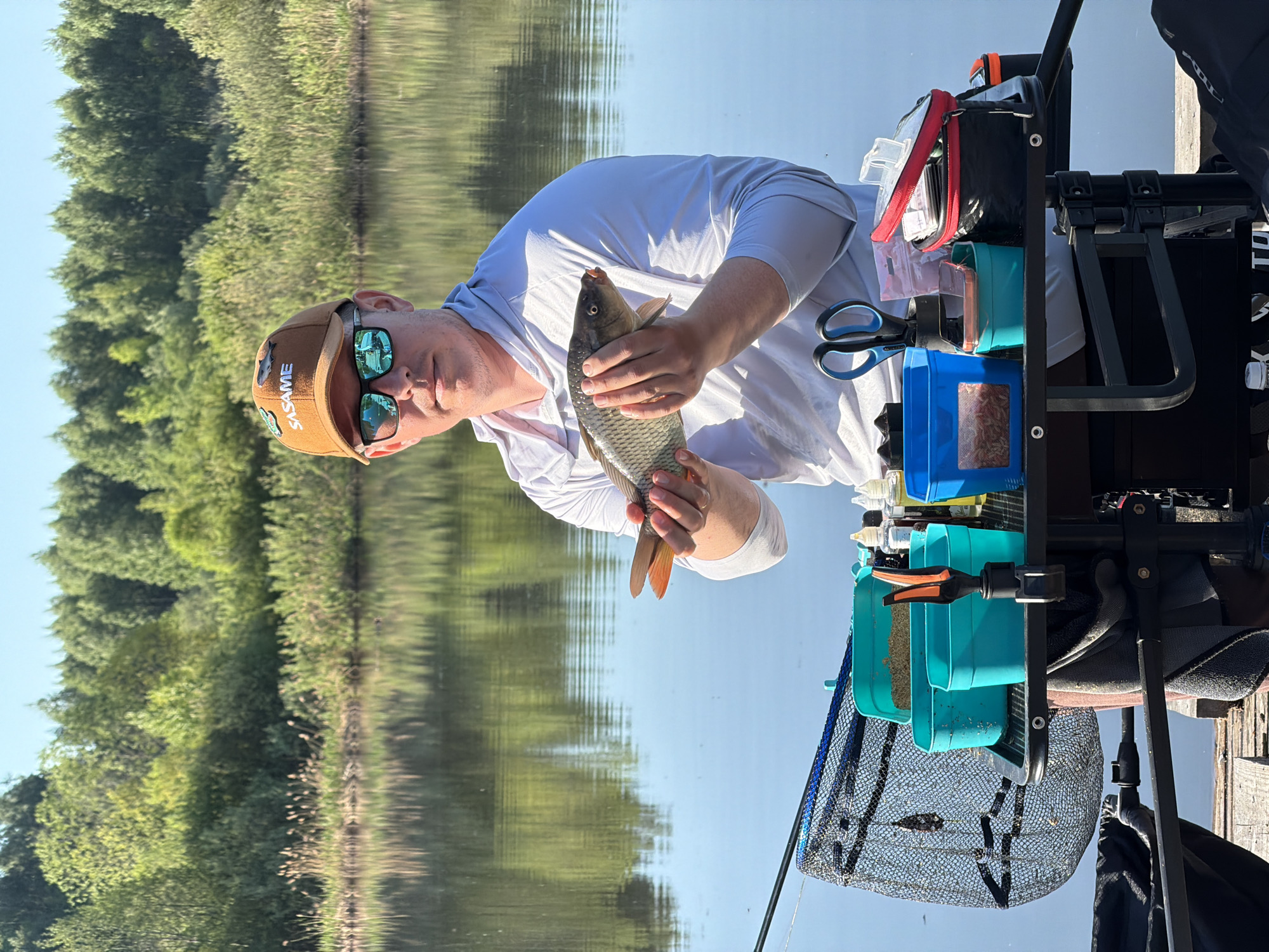 A man wearing a brown cap and sunglasses holds a fish in his hands next to a fishing station. The location appears to be outdoors next to a body of water, with trees visible in the background. The image is related to fishing. The city of Belaya Gora, Russia, cannot be confirmed from the image.