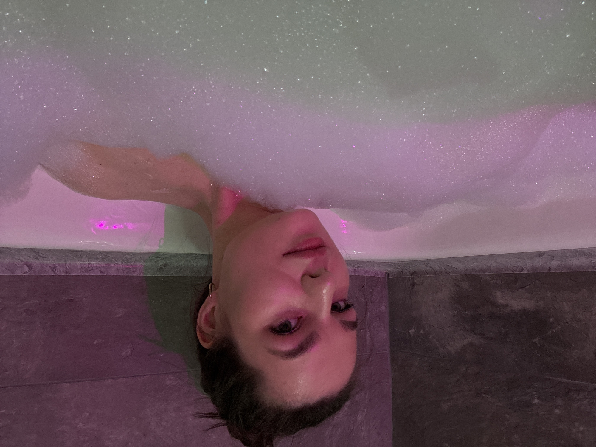 A young woman is submerged in a bubble bath in a bathroom with dark gray tiling. The scene is illuminated by pink lighting. Her face is visible, and she appears relaxed. The location is presumably a bathroom in Chaikovsky, Russia.