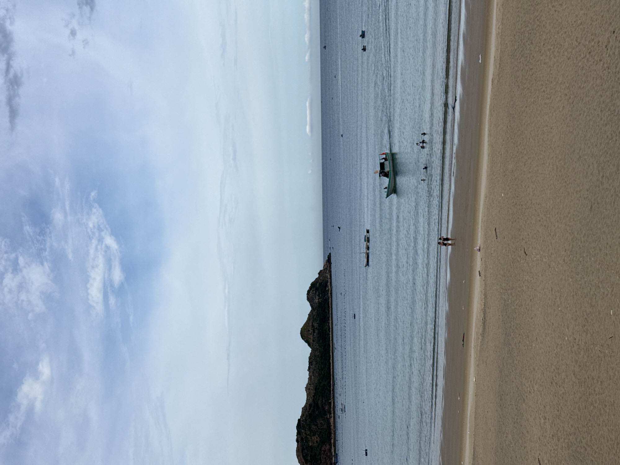 The image shows a beach with a calm ocean and a cloudy sky. In the foreground, there is a sandy beach with a few pieces of debris. Two people are walking along the shoreline. In the middle ground, the ocean is blue and there are several boats. One boat in the center is a small fishing boat with people on board. There are also a few swimmers in the water. In the background, there is a large, rocky island covered in vegetation. The horizon line is visible, separating the sea from the sky. The overall scene depicts a peaceful coastal environment.