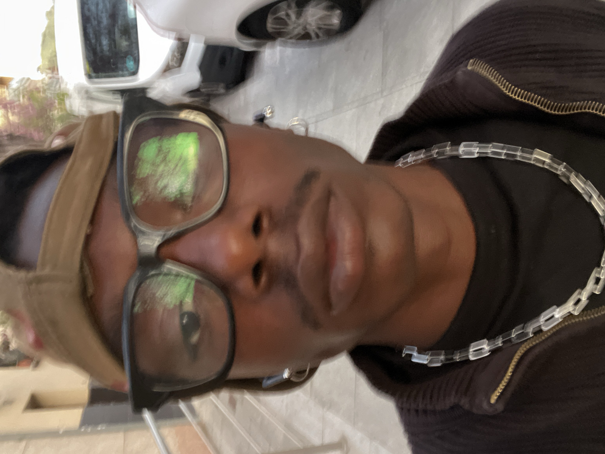 A selfie taken by a person with dark skin. The person is wearing glasses, a cap, a dark-colored zipped jacket, and a transparent necklace. The reflection in the glasses suggests trees or vegetation. A white car is visible in the background. Based on the architectural style and the presence of palm trees, the location appears to be Barcelona, Spain.