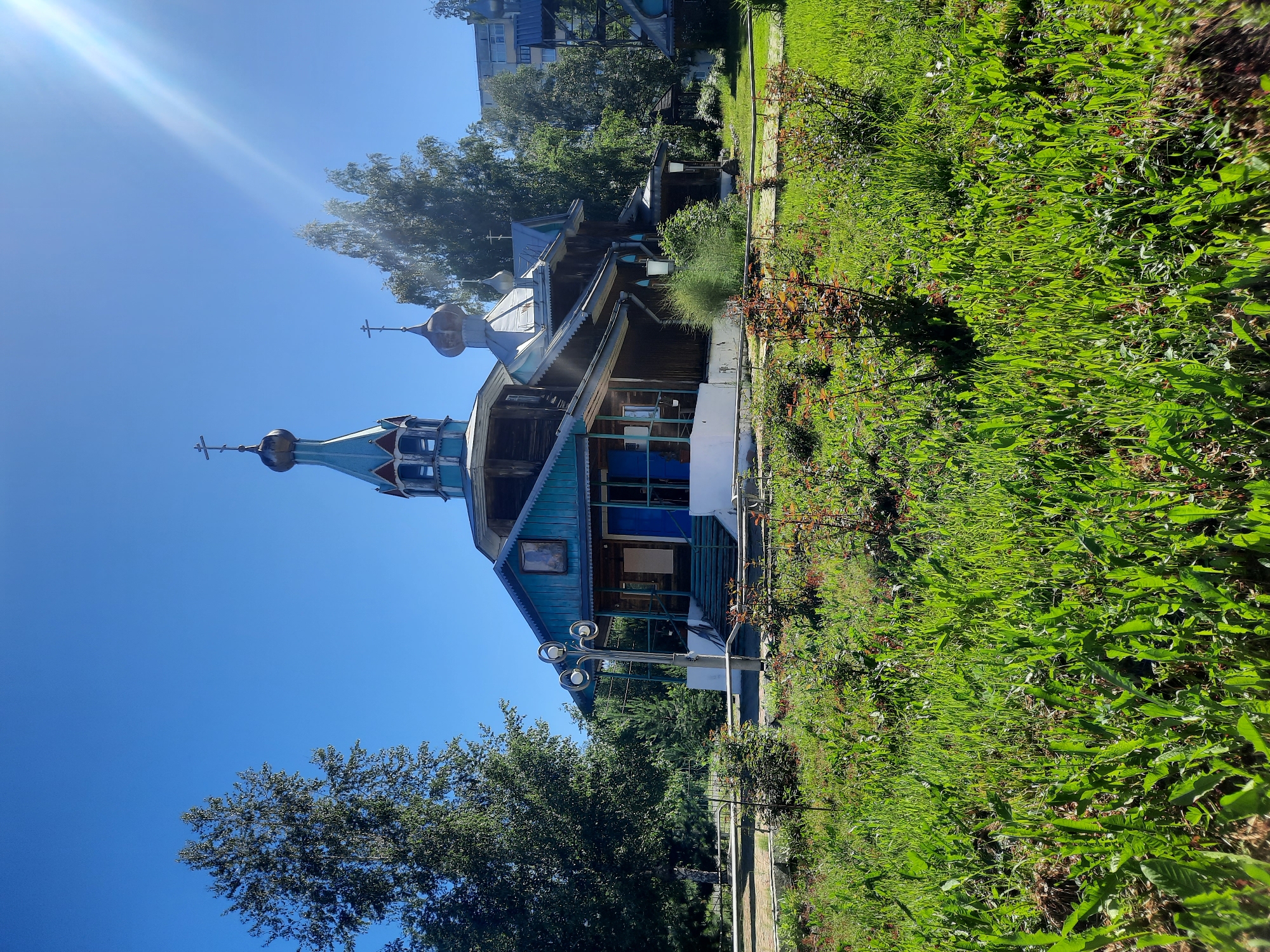 The image shows an Orthodox church with a wooden structure and blue accents. The church has multiple domes, each topped with a cross. In the foreground, there is a well-maintained lawn with small bushes and flowering plants. A pathway made of stone or concrete is visible in the middle ground, leading towards the church. A street lamp with multiple light fixtures stands to the left of the pathway. Behind the church, there are trees and a residential building. The sky is clear and blue with a bright sun, casting a lens flare in the upper right corner. The scene is set in a serene outdoor environment, possibly a park or a churchyard, in the city of Zelenogorsk, Russia. There are no people visible in the image.