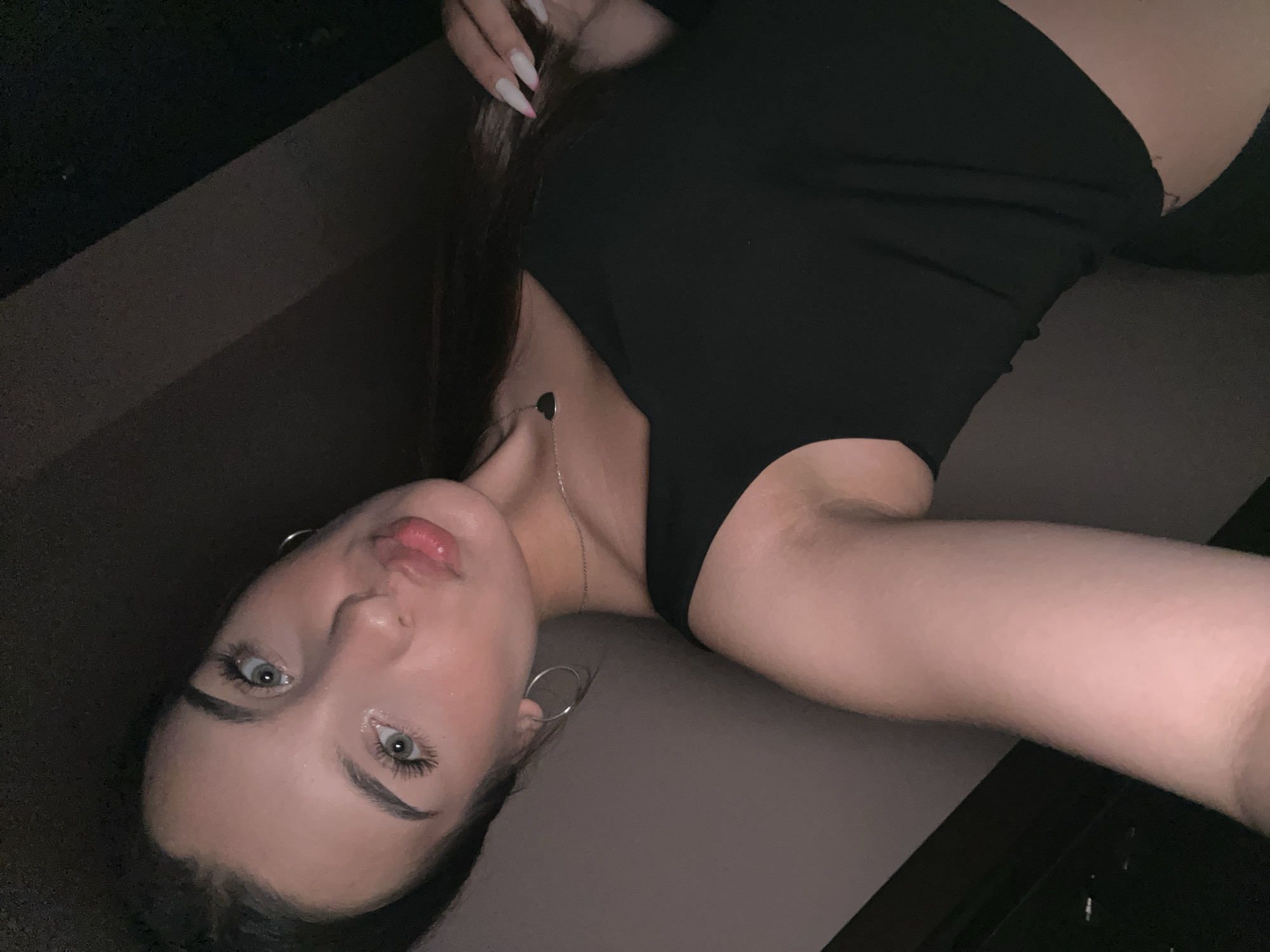 This is a selfie of a young woman with light skin, dark hair, and blue eyes. She is wearing a black off-the-shoulder top and a silver necklace with a heart pendant. Her lips are slightly parted and pursed. Her right arm is visible, showing long fingernails with white and pink tips. The background is dark and appears to be an indoor setting, possibly a room with a wall and a dark surface. The lighting is dim, with the subject's face illuminated. The location is not specified in the image.