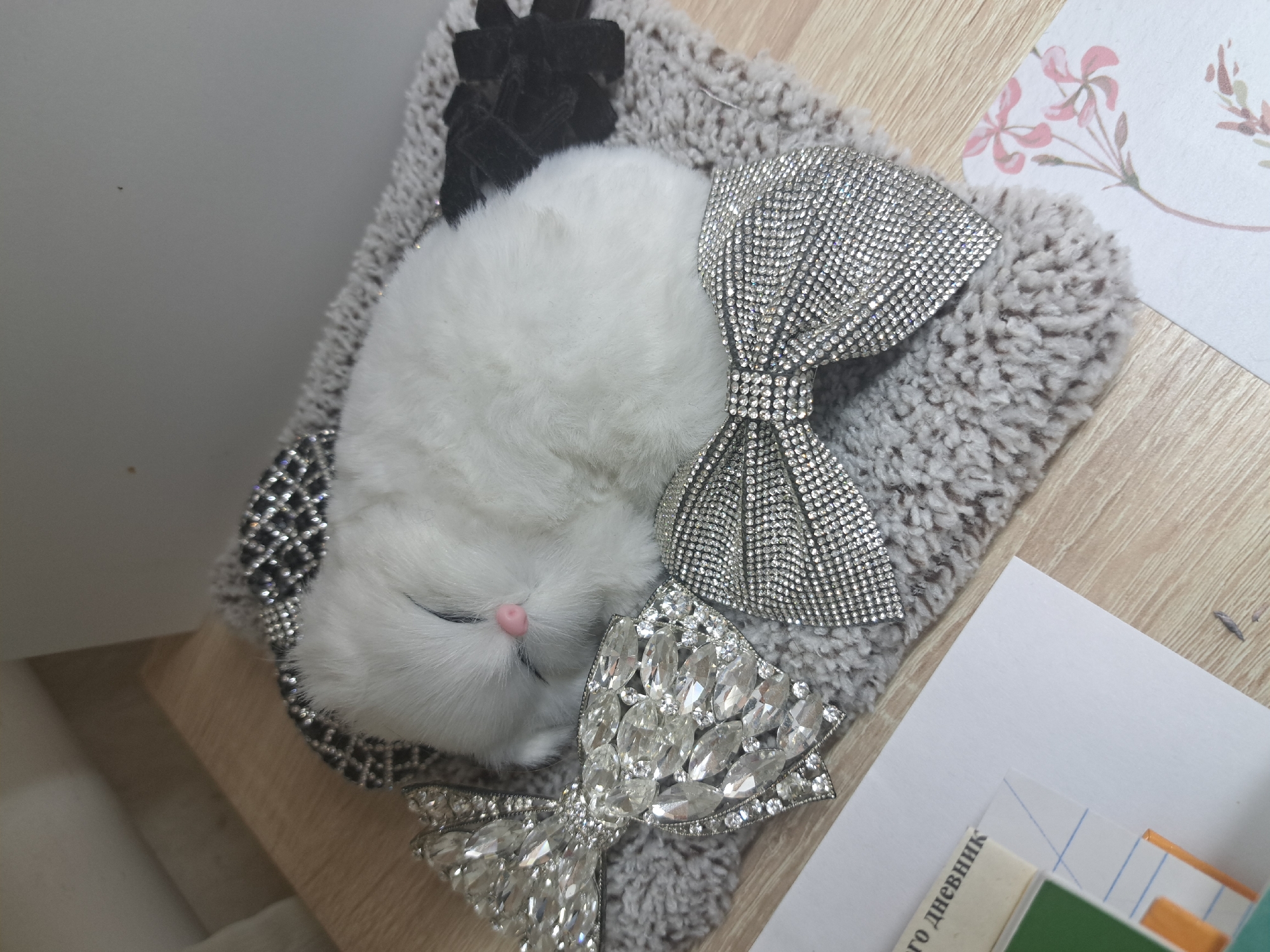 A white, fluffy animal toy with a pink nose is lying on a gray, textured cushion. The cushion is decorated with three bows: one black velvet bow, one silver bow covered in small crystals, and one larger silver bow covered in larger, more irregularly shaped crystals. The cushion is placed on a light-colored wooden surface. To the right of the cushion, there is a white paper with a faint floral design and a stack of papers with text in Russian ("-го дневник"). The location is unknown.