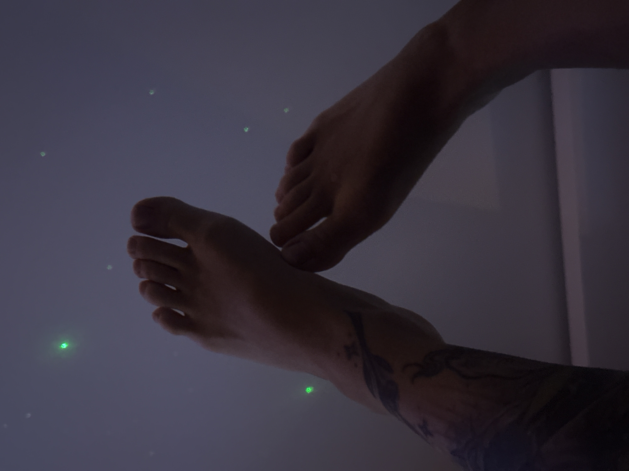 The image shows two human feet against a dark, speckled background, possibly a ceiling or wall with decorative lights. One foot is positioned closer to the viewer, showing the toes and top of the foot. The other foot is slightly further away and angled. A tattoo is visible on the lower leg and ankle of the closer foot. The background features several small, blurred green lights, resembling stars. There is no discernible indication of a specific location in Moscow, Russia. The scene is static and focuses on the feet and background lights.