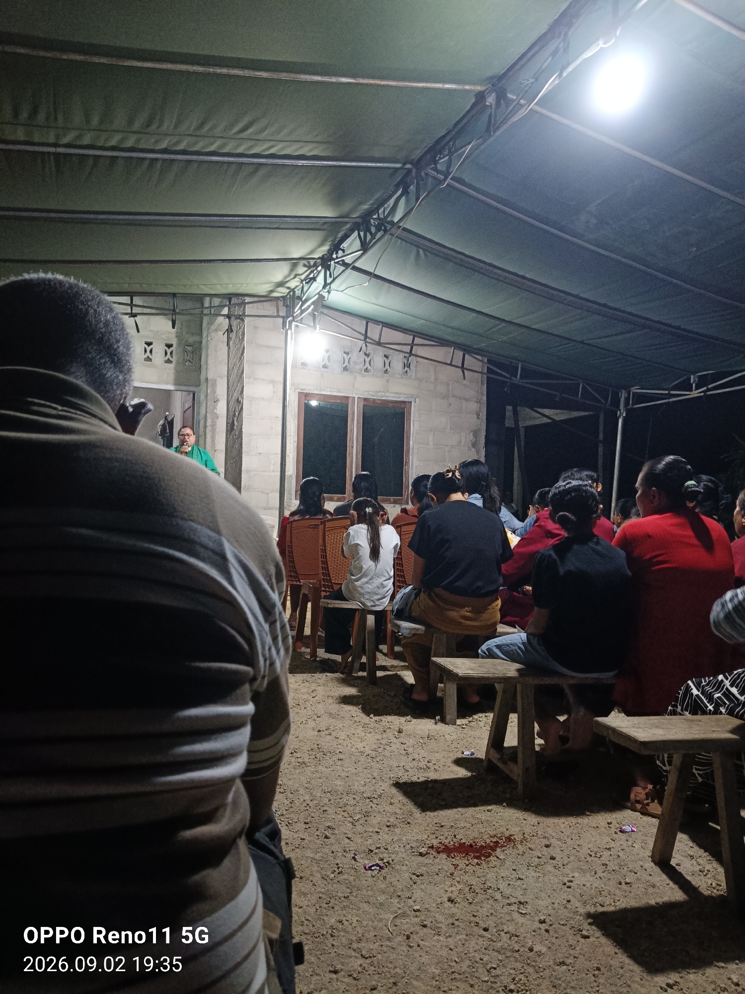 The image depicts an outdoor gathering at night, possibly in Otan, Indonesia, beneath a green canopy supported by metal poles and a wooden structure. The ground is covered with gravel and dirt. In the foreground, the back of a person wearing a striped shirt is visible. In the mid-ground, a group of people are seated on wooden stools, facing a woman in a green shirt who is speaking into a microphone. The building behind them is constructed with concrete blocks and has windows. The scene is illuminated by artificial lighting. There is a small stain on the ground in the mid-ground that appears to be blood. The timestamp on the image indicates it was taken on September 2, 2026, at 19:35.