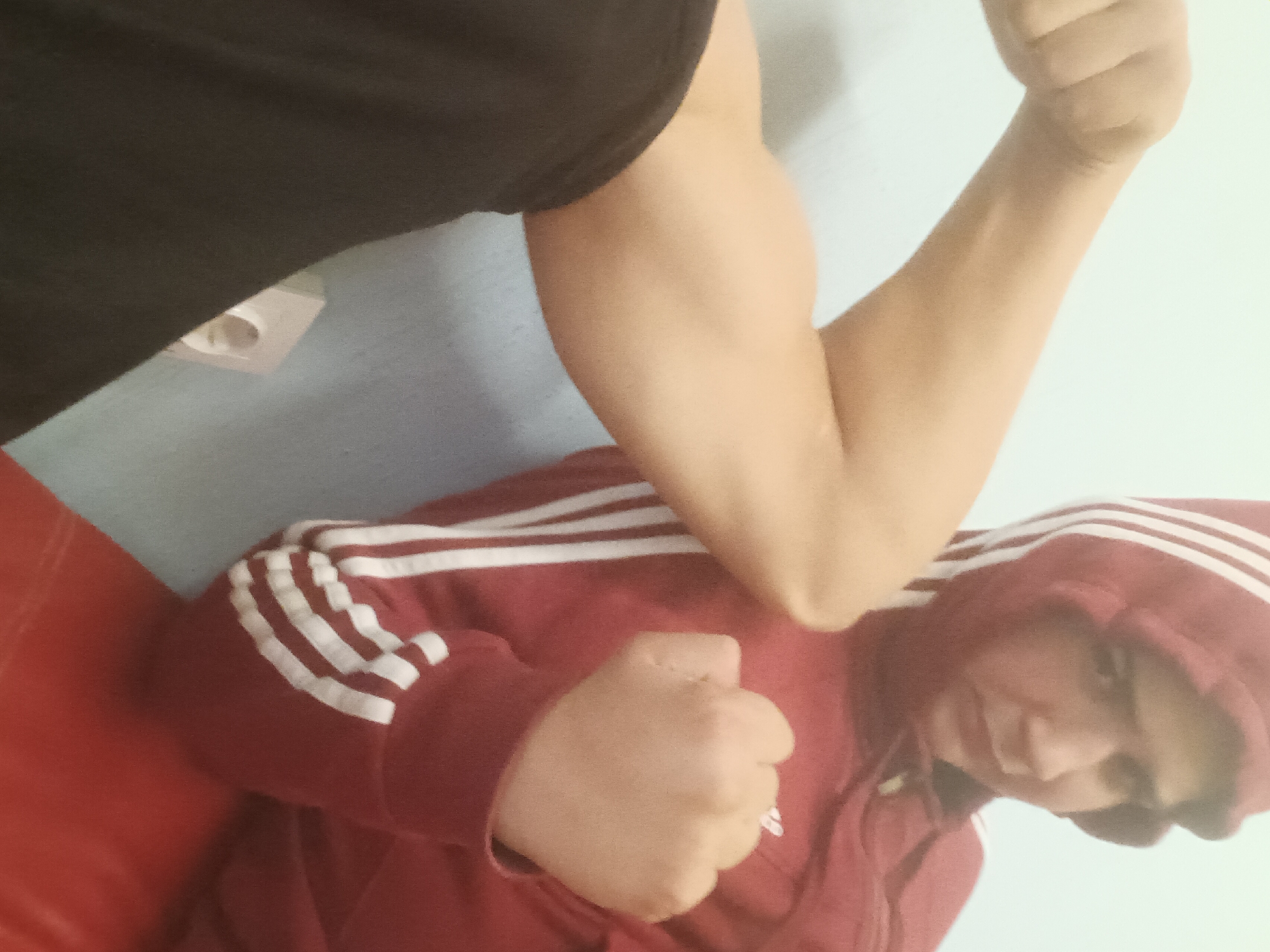 A person wearing a red hoodie with white stripes is lying on a red surface with a person applying an arm lock. The person applying the lock is wearing a black t-shirt and the arm is pressing on the other person's neck. They are on a light blue wall with a white electrical outlet. The city and specific location are unknown.