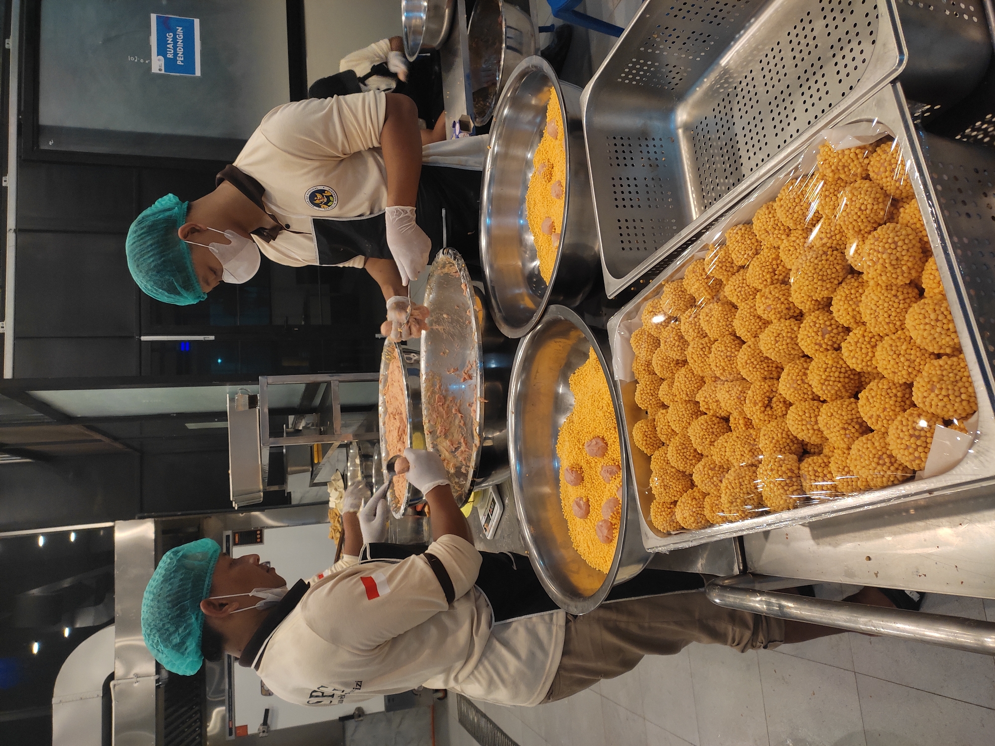The image shows two men wearing hairnets and face masks, working in a commercial kitchen. They are both wearing similar light-colored shirts with dark aprons. The man on the left is holding a scooper and a bowl of ground meat mixture, while the man on the right is scooping the meat mixture into a bowl. In the foreground, there is a large metal tray filled with small, round, yellow-orange food items, some of which appear to be coated in small seeds or granules. Next to this tray is another metal container, possibly for storage or transport. In the background, there are stainless steel countertops, cooking equipment, and a sign that says "RUANG PENDINGIN" which translates to "REFRIGERATED ROOM" in English. This scene appears to be taking place in a food preparation area.