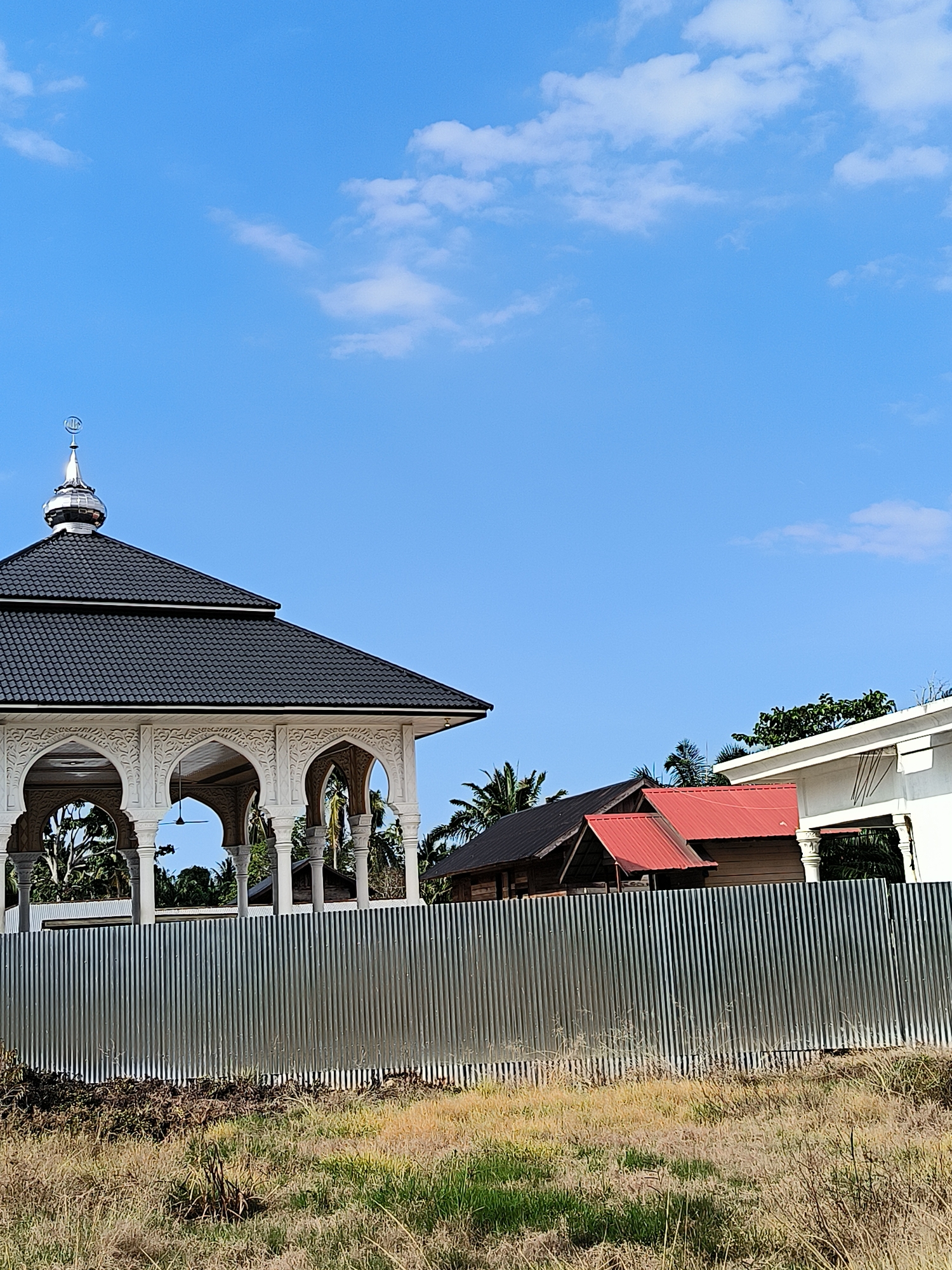 The image shows a building, likely a mosque or Islamic structure, in Aluebutuha, Indonesia. The building has a black tiled roof, white ornate columns and arches, and a silver dome with Islamic crescent symbol on top. In the background, there are two other buildings, one with a red roof and the other with a dark roof. A corrugated metal fence runs along the front of the property, and there is a field of dry grass in the foreground. The sky is blue with scattered white clouds. There are no people visible in the image.