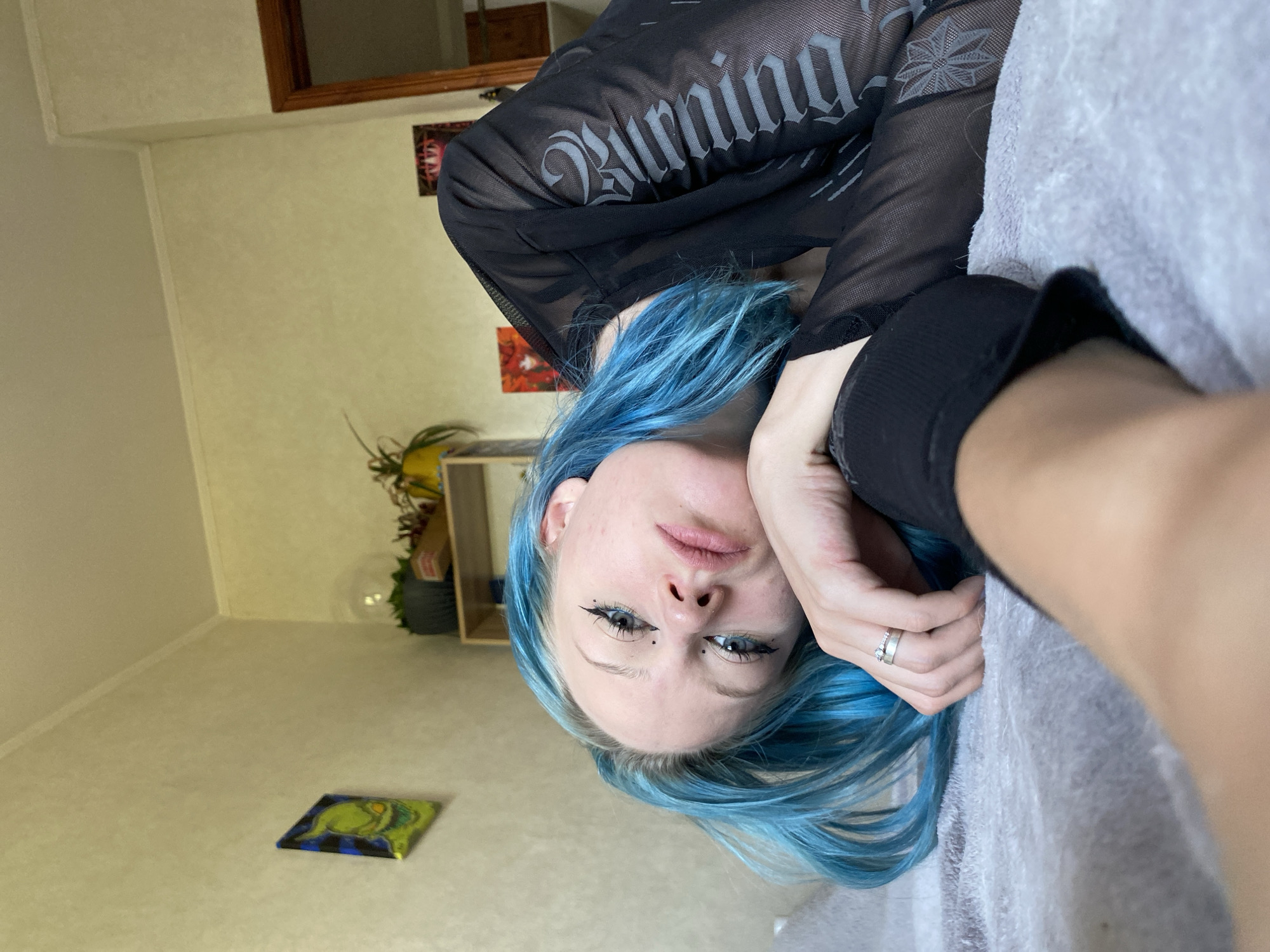 A young woman with bright blue hair is lying on a bed, resting her head on her arm. She is wearing a sheer black long-sleeved shirt with a gothic-style pattern. Her eyes are open, and she is looking towards the camera with a neutral expression. She has dramatic winged eyeliner and small black dots under her eyes. A silver ring is visible on her right hand.
The room appears to be a bedroom. The walls are a light, textured color. On the wall to the left, a small, colorful painting depicting a cartoonish, green character is hanging. To the right, there is a wooden shelf with potted plants and a glass globe. Further to the right, a doorway with a wooden frame is visible, and a small framed picture hangs on the wall near it. The bed is covered with a light gray textured blanket. The lighting suggests it is daytime, with light coming from a window not visible in the frame. The overall impression is a casual, indoor setting.