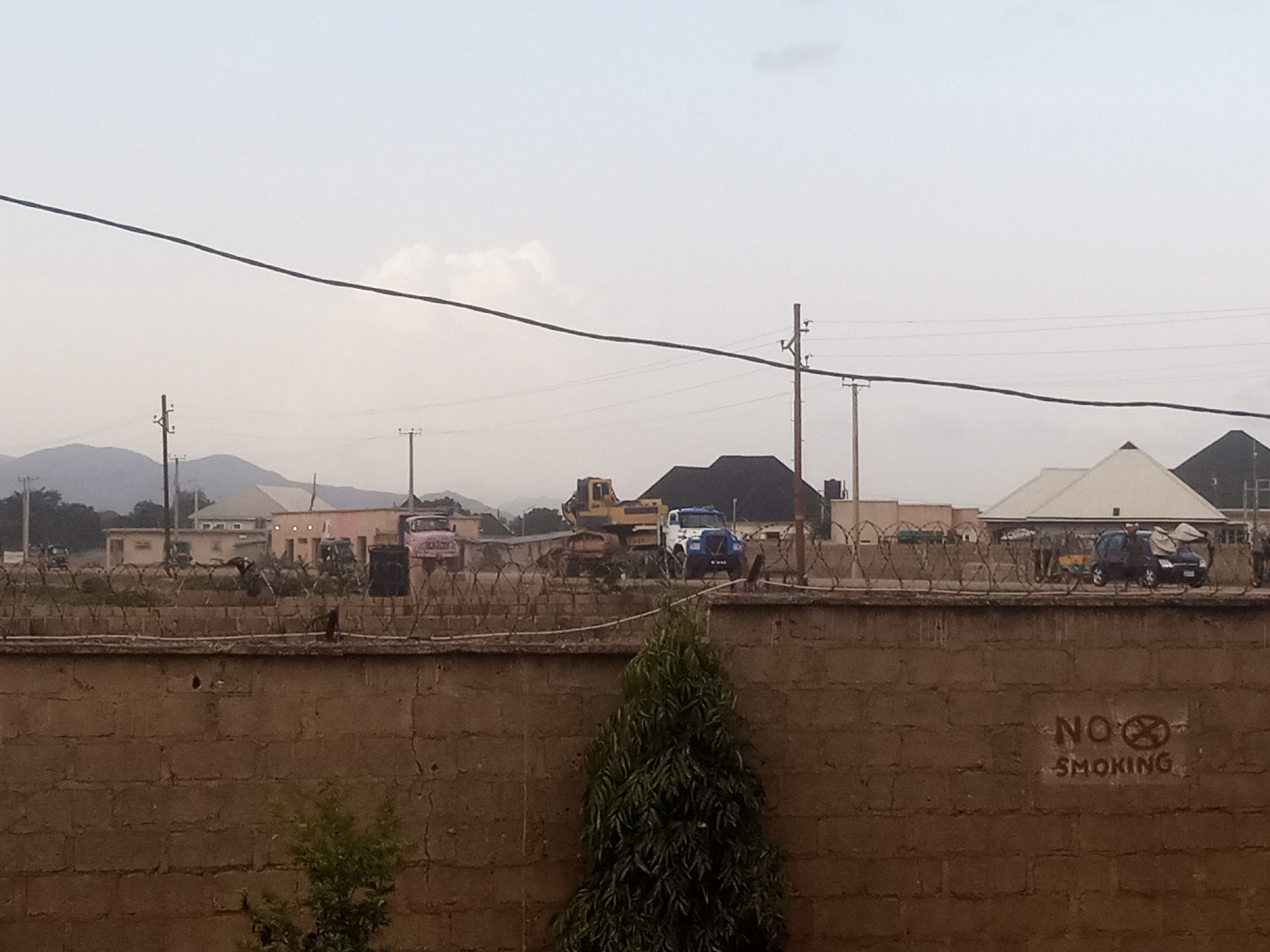 This media file shows an outdoor scene with a brick wall in the foreground, topped with barbed wire. Behind the wall, there are several buildings, including some with distinctive roof shapes, possibly indicating a commercial or residential area. A yellow excavator and various vehicles, including trucks and what appears to be a pink truck, are visible. The sky is overcast, suggesting a cloudy day. Power lines and poles are prominent across the scene, indicating infrastructure.
On the brick wall in the foreground, there is visible text that reads "NO SMOKING" with a circular symbol crossed out over the "O" in "NO". The overall impression is of an industrial or construction site, or perhaps a secure compound, given the barbed wire and the presence of heavy machinery. The lighting suggests it could be daytime, but the overcast sky diffuses the light. No specific emotions are conveyed, but the scene appears to be a static view of an environment rather than a dynamic event. The background shows a gentle slope of hills or mountains.