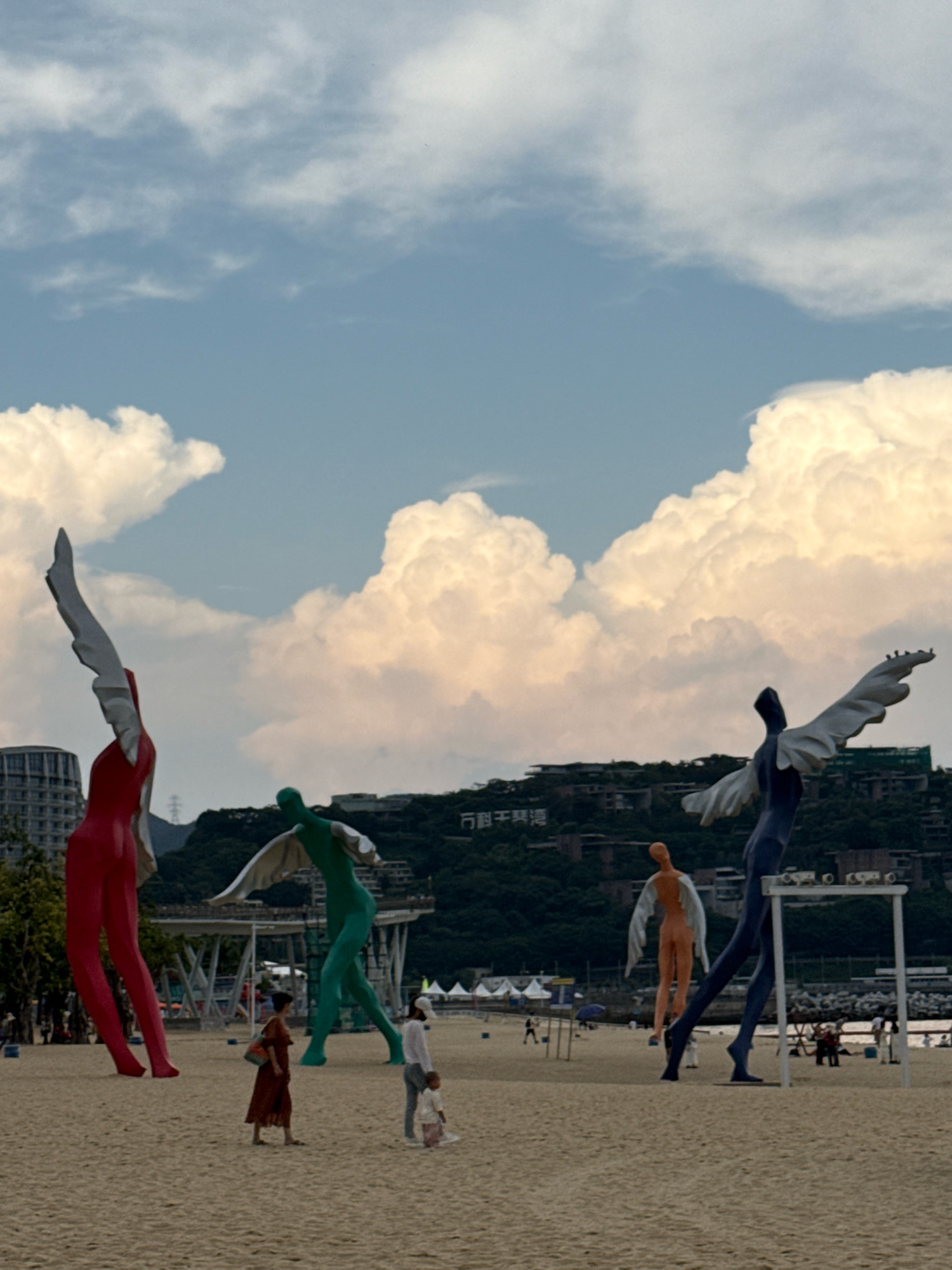 This image depicts a beach scene in Yantian District, China, with a focus on several large, abstract human-like sculptures. The sculptures, varying in color from red, green, blue, and orange, feature wings, suggesting an angelic or mythical theme. They are positioned on a sandy beach with the ocean visible in the background, along with a treed hillside with buildings and a sign in Chinese characters. The sky is partly cloudy with dramatic, puffy clouds. Several people are visible on the beach, including two adults and a child in the foreground, as well as other figures in the mid-ground. The overall impression is of an outdoor art installation on a public beach.