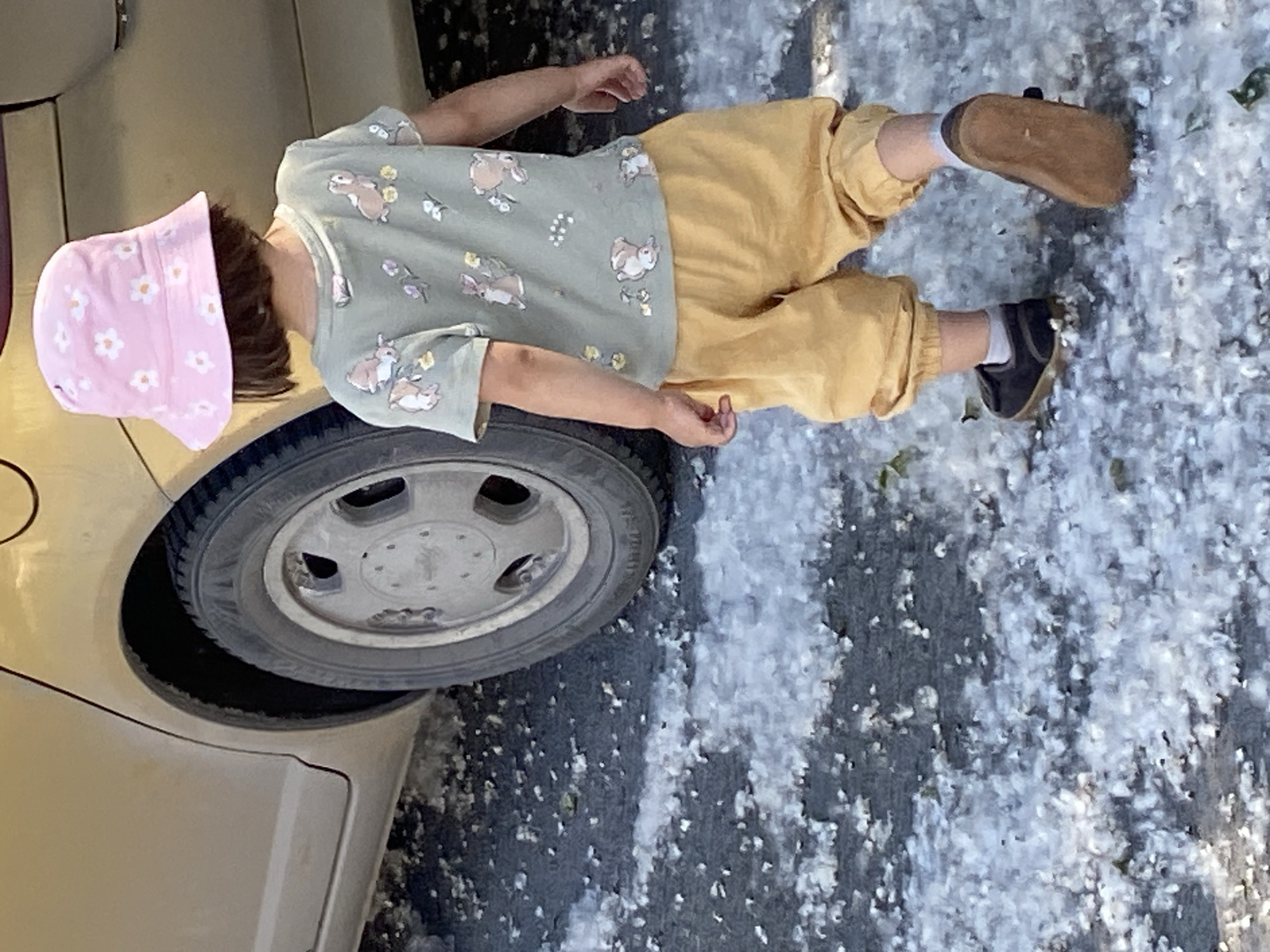 An outdoor, daytime image captures a young child, viewed from behind, walking on a dark paved surface largely covered with white, fluffy material, consistent with cottonwood seeds. The child is positioned to the right of the rear section of a light-colored sedan. The child wears a pink bucket hat adorned with white floral patterns, a light green short-sleeved t-shirt featuring multiple cartoon-style rabbits and small floral designs, and loose-fitting mustard yellow pants cuffed at the ankles. Dark shoes with light brown soles and white socks are visible on their feet, with the right foot partially lifted, showing the sole. The car's rear quarter panel and a wheel with a silver multi-spoke hubcap are visible on the left side of the frame. No specific visual identifiers within the image confirm the location as Tyumen, Russia.