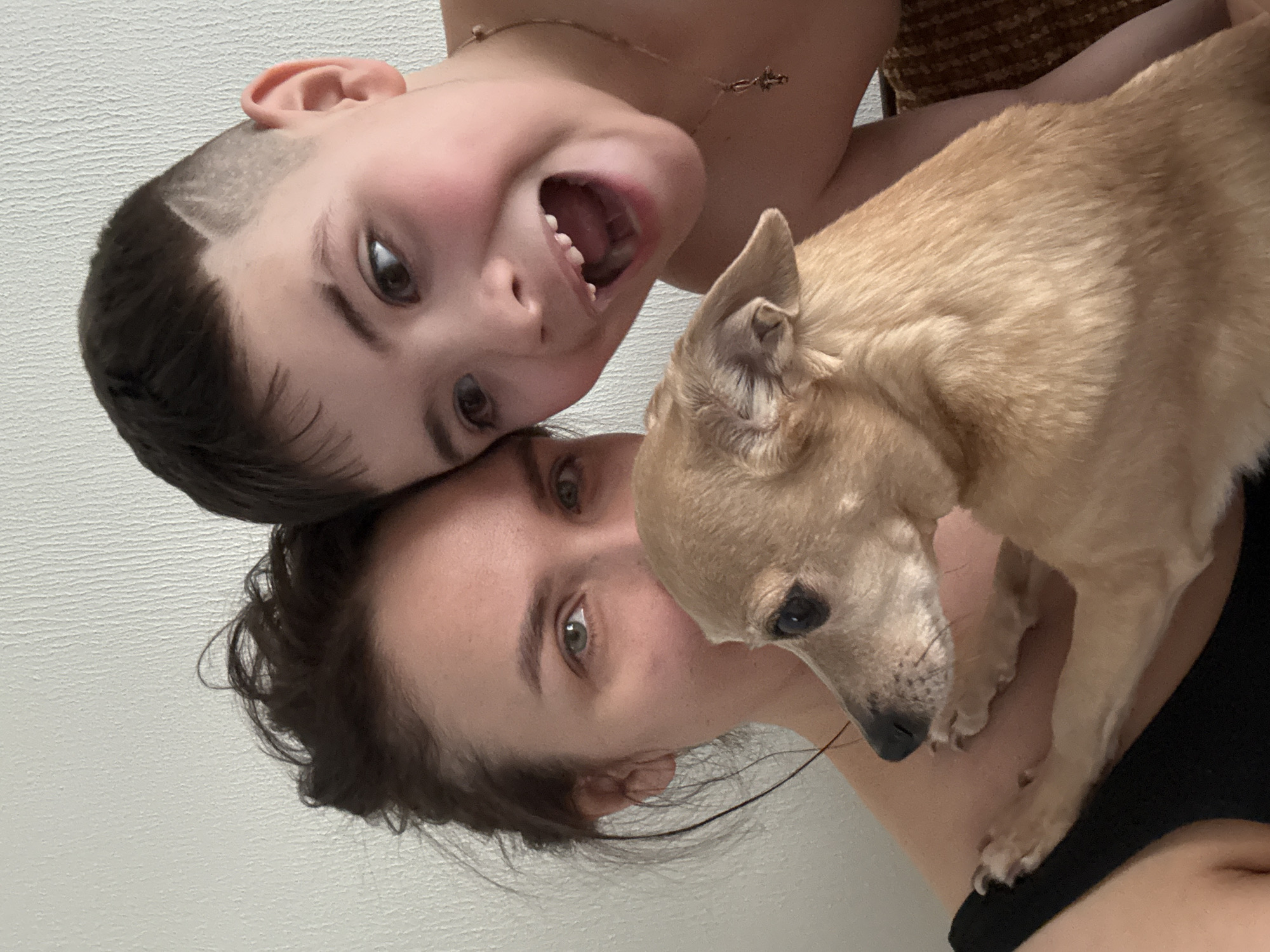 In an indoor setting in the city of Chaikovsky, Russia, a portrait features an adult female, a male child, and a small dog. The adult female, positioned on the left, has dark hair pulled back and green eyes, with her face partially obscured by the dog. She wears a dark top. A male child, with short dark hair featuring a shaved design on the right temple, is on the right, shirtless, wearing a gold-colored necklace with a cross pendant. He has his mouth wide open, displaying teeth, and is looking towards the camera with an expressive facial gesture. A small, short-haired, light-tan colored dog is held by the female, positioned centrally and facing left in profile. The dog's paws are visible on the woman's arm. The background is a light-colored, subtly textured wall, with a small portion of a brown, textured surface visible on the far right.
