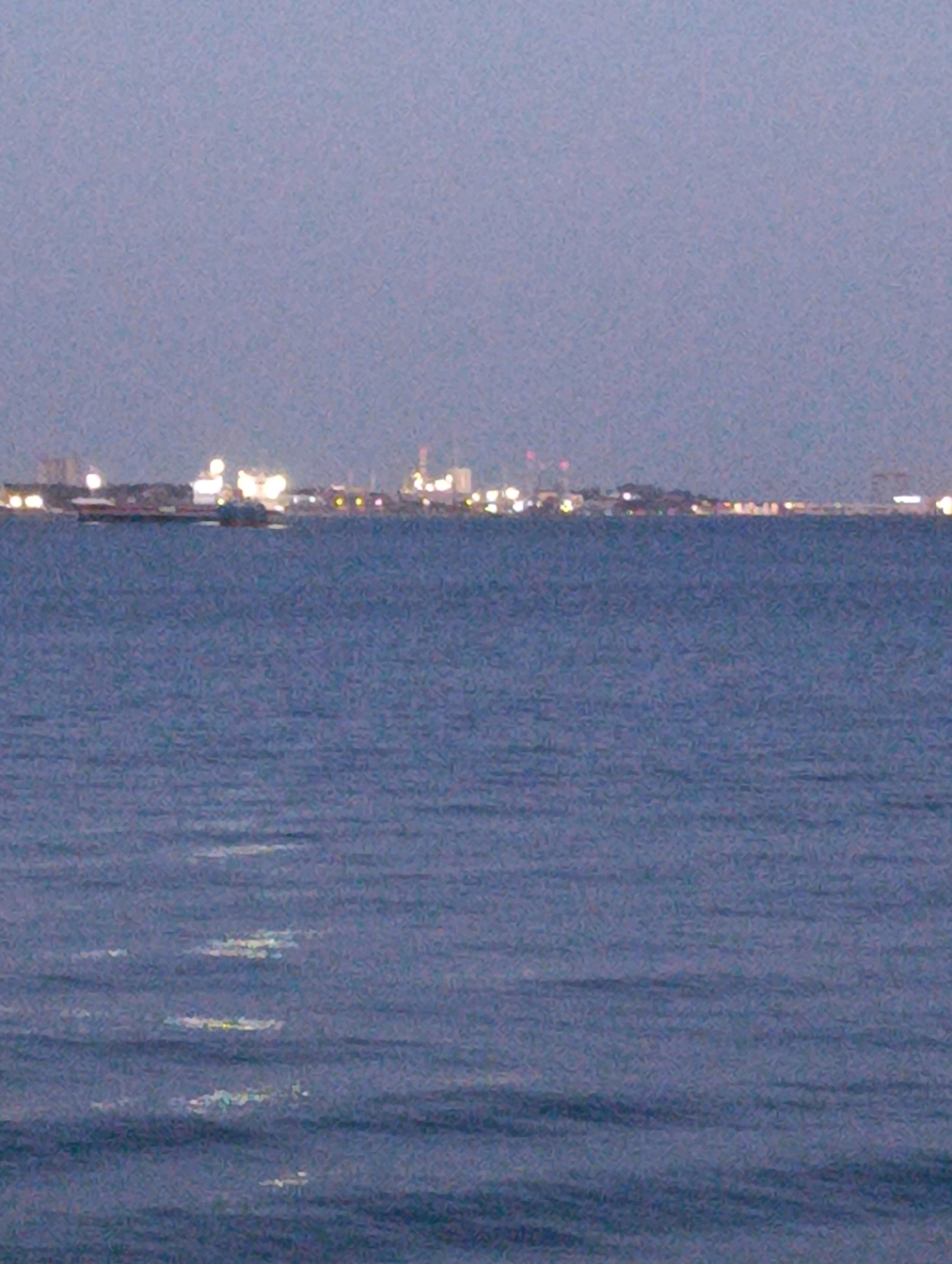 The image depicts a hazy, twilight view across a body of water towards a distant, illuminated cityscape. The water in the foreground is a deep blue with subtle ripples, reflecting faint, elongated streaks of light. In the middle ground, a large, dark vessel, possibly a ship or barge, is silhouetted against the horizon. Beyond the water, the cityscape is characterized by a continuous line of buildings and structures, many of which are illuminated by numerous lights, suggesting an urban or industrial area. The overall atmosphere is subdued, with a low-light quality typical of dusk or dawn. The location is identified as the city of Setúbal, Portugal.