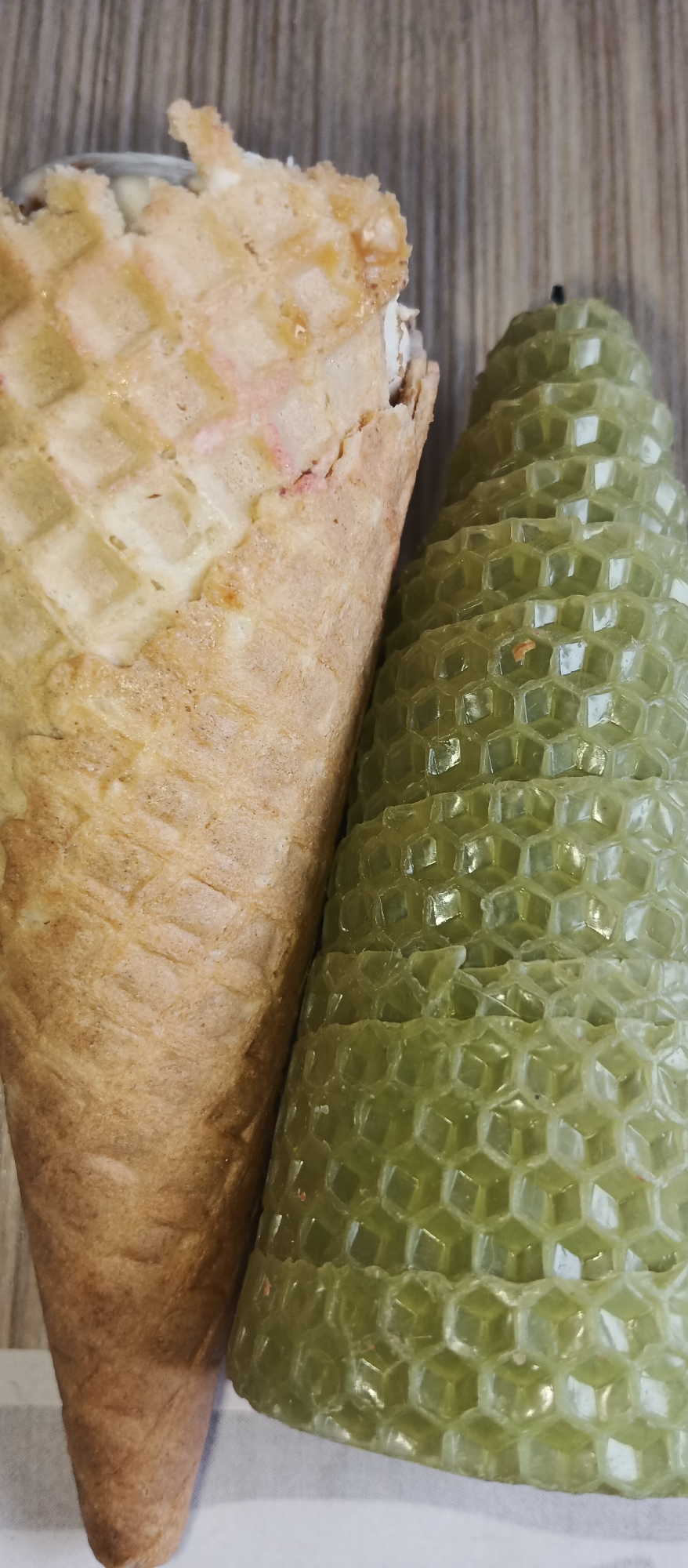 A close-up, vertical photograph displays two distinct objects positioned side-by-side. On the left is a light brown waffle cone, likely an ice cream cone, characterized by its prominent grid-like, square waffle pattern. The top edge of the cone is slightly irregular, and some light-colored, possibly pinkish or white, filling is visible within its opening.
Adjacent to the waffle cone, on the right side of the frame, is a pale green, conical candle. This candle has a distinctive, textured surface composed entirely of interconnected hexagonal cells, resembling a honeycomb or the scales of a pinecone. A small black wick is visible at the very top of the candle.
The background behind the objects transitions from a light brown surface with a vertical wood grain pattern at the top to a plain white or light-colored surface at the bottom. The objects are static, with no activity or event taking place. The lighting is even, emphasizing the textures of both the waffle cone and the candle. There is no visible text in the image.