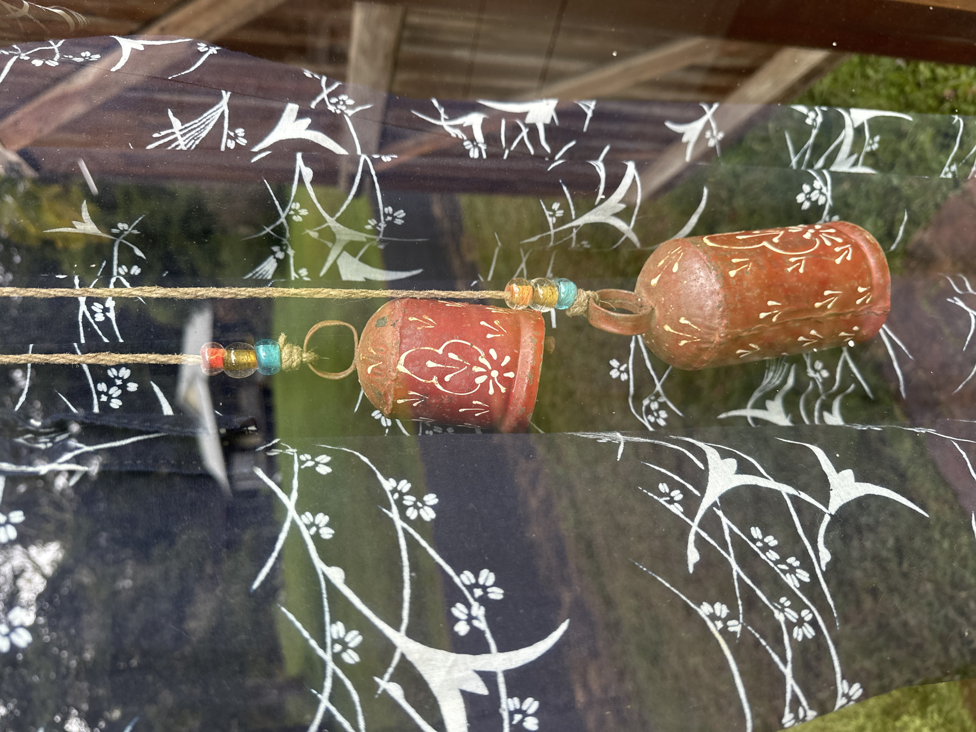 The image shows two rustic, bell-shaped wind chimes hanging from a jute string, adorned with colorful beads. The wind chimes are decorated with white, stylized floral patterns and appear to be made of metal with an aged patina. They are suspended in front of a dark fabric with a white, bird and branch motif. Through the fabric and what appears to be a window, a blurred outdoor scene is visible, featuring green grass, trees, and a hint of a wooden structure. The overall impression is a decorative piece set against an outdoor backdrop, with no people or specific location markers in Thora, Australia evident.