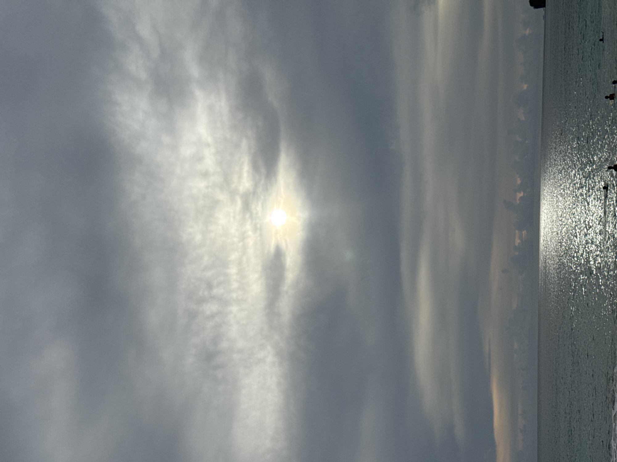 The image depicts a serene ocean scene with a cloudy sky and the sun partially visible. The sky is filled with varying shades of gray clouds, with the sun creating a bright, diffused glow through a thin patch of clouds, suggesting a hazy or overcast day. The water reflects the sky's muted light, creating a shimmering surface with streaks of light.
In the distance, along the right side of the image, a faint shoreline or landmass can be observed. Scattered along the edge of the water and in the shallows, there are a few indistinct silhouettes of people. Their presence suggests they are either wading, swimming, or standing at the water's edge. The overall mood is peaceful and tranquil, with the diffused sunlight and calm water contributing to a sense of quietude. The time of day appears to be either early morning or late afternoon, given the low angle and diffused quality of the sunlight. There is no visible text in the image. The scene evokes a sense of calm and natural beauty, typical of a beach at dawn or dusk.