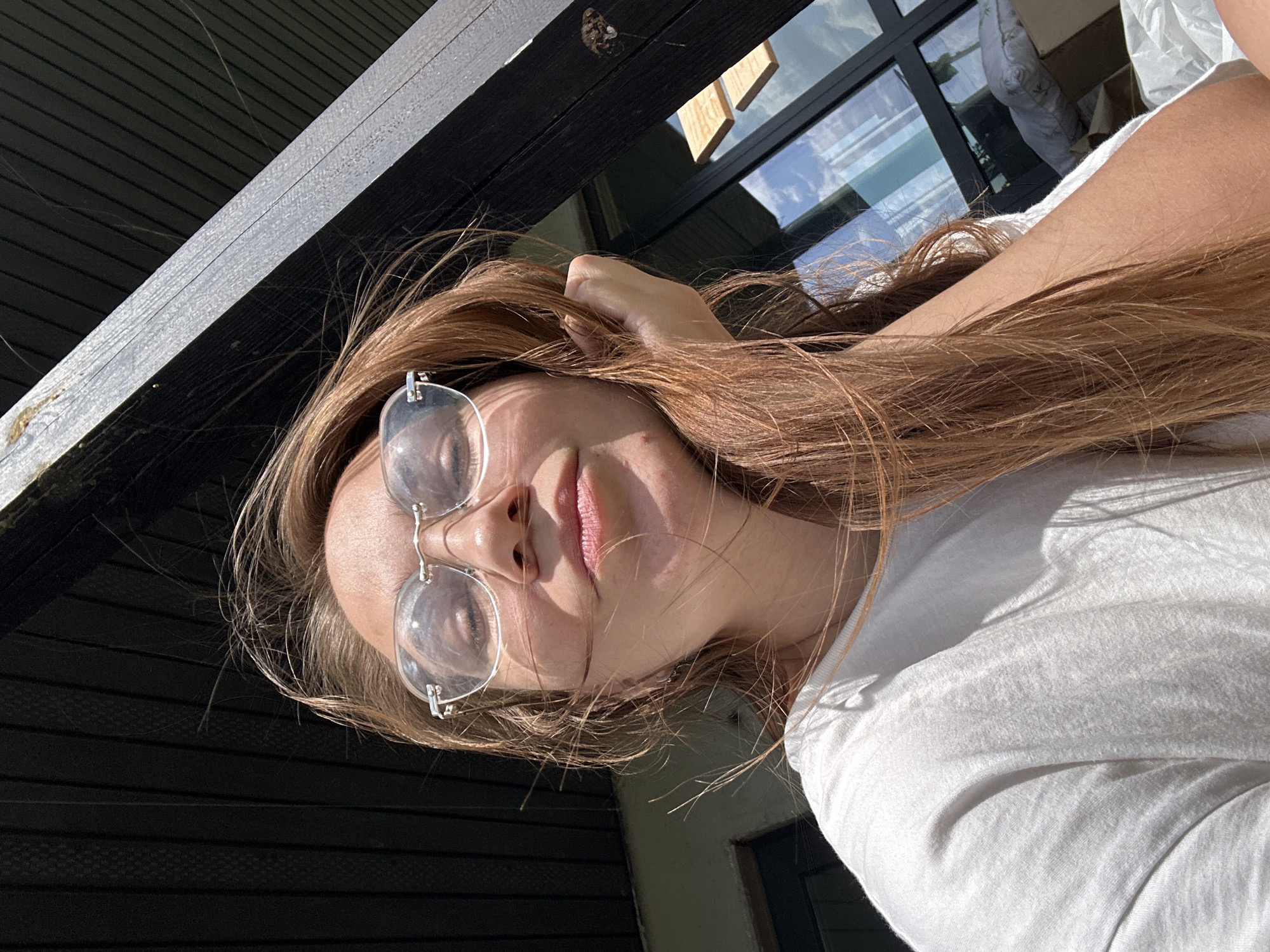 A young woman with long, light brown hair is facing the camera and slightly angled to the left. She is wearing silver-rimmed glasses with circular lenses. She has fair skin, a small nose, and pink lips. She is wearing a white t-shirt. Her left hand is raised and appears to be touching her hair. The background includes a dark wooden structure and a glass window reflecting the sky. The location is not discernible from the image.