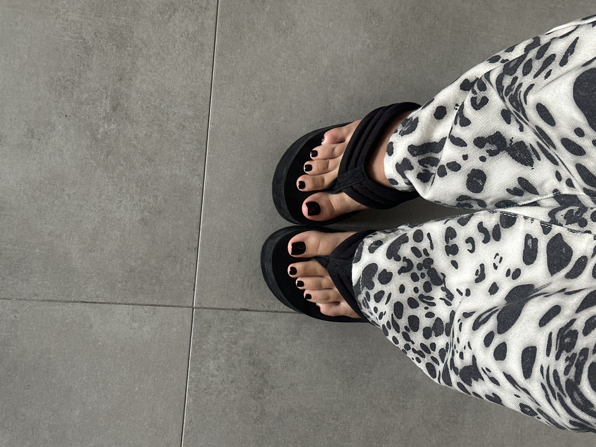 The image shows a pair of feet wearing black platform flip-flops and leopard-print pants, standing on a gray tiled floor. The toes are painted with dark nail polish. The lighting is natural, casting soft shadows. The scene is an indoor environment with no visible city landmarks or specific locations in Choeng Thale, Thailand.