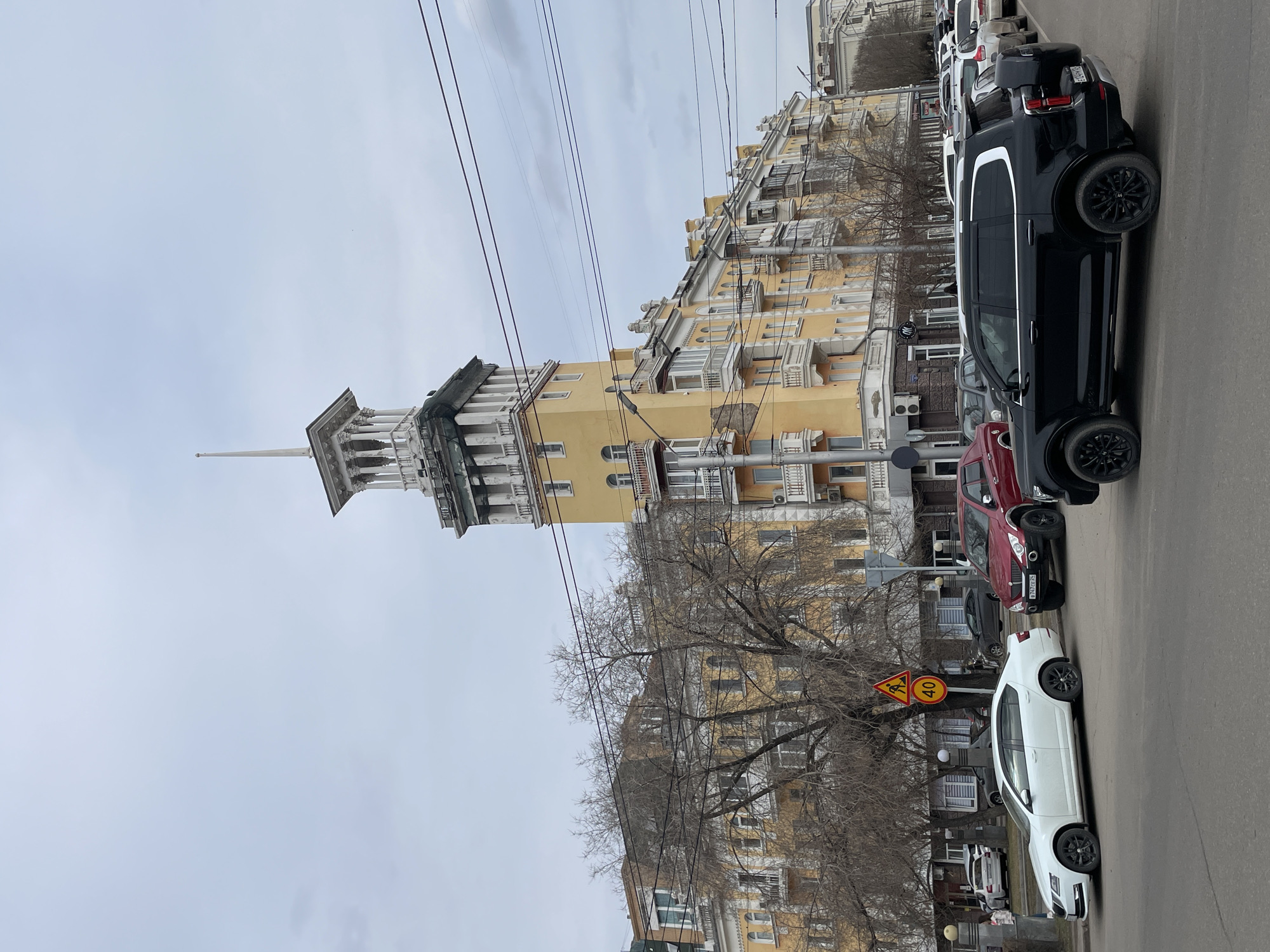 The image depicts a view of Krasnoyarsk, Russia, showcasing a prominent yellow Stalinist-era building with a distinctive tower. The building is a defining architectural feature. Several cars are parked along the street in front of the building, including a white sedan, a red SUV, and a black van. A road sign indicating a speed limit of 40 km/h and warning of a pedestrian crossing is visible. Bare trees line the street, and power lines stretch across the sky. The weather appears overcast. The image is taken from street level.