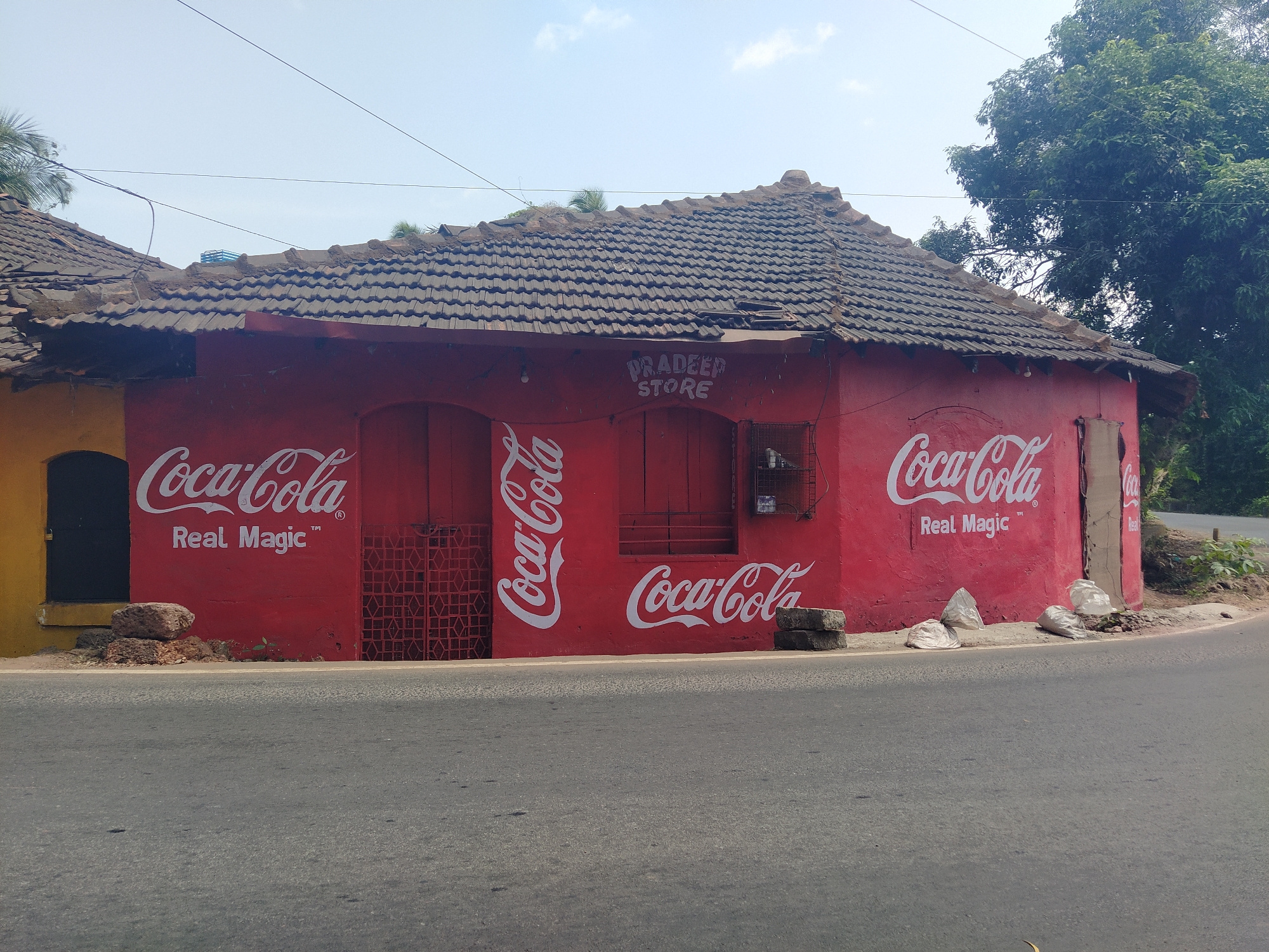 The image shows a red-painted building in Moira, India, prominently adorned with Coca-Cola advertisements. The building, identifiable as "Pradeep Store" from signage above a window, features a tiled roof with some visible damage. "Coca-Cola" and "Real Magic" are painted in white on multiple sides of the structure. The building abuts a paved road. The environment includes trees and other vegetation in the background. Debris such as rocks and plastic bags are visible along the base of the building. A small yellow building is on the left side.