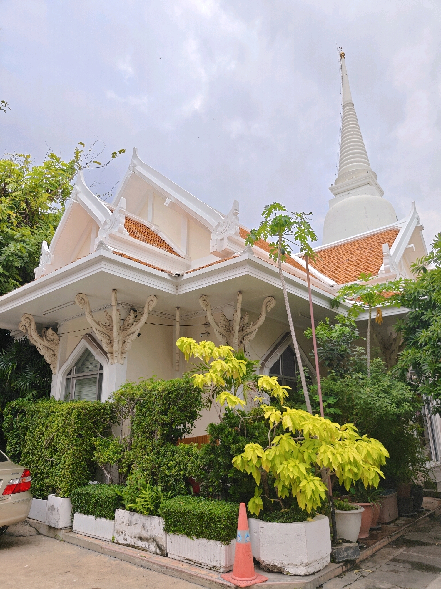 The image shows a Buddhist temple with a white stupa in the background. The temple has traditional Thai architecture with a white ornate facade and a terracotta tiled roof. The building is surrounded by lush greenery, including various trees and bushes in pots and planters. A beige car is parked on the left side of the image. An orange traffic cone is placed in front of a planter on the right. The sky is overcast. This temple appears to be Wat Suthat Thepphawararam, a significant Buddhist temple located in Bangkok, Thailand.