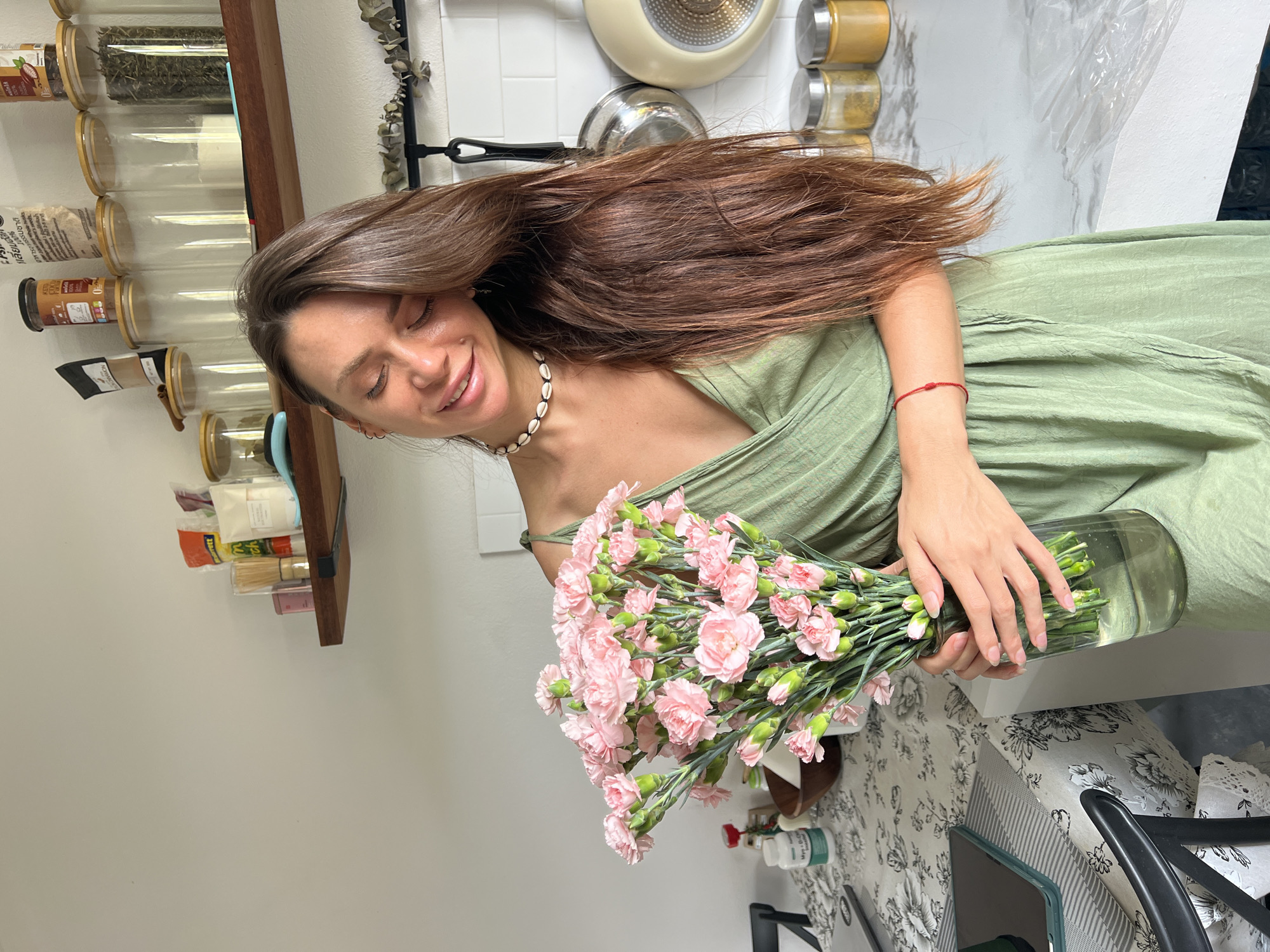 The image depicts a woman indoors, likely in a residential kitchen, possibly in บ้านบางรักษ์, Thailand. She is holding a large bouquet of pink carnations in a clear glass vase. The woman has long brown hair and is wearing a light green dress. She is also wearing a shell necklace and a red string bracelet. The kitchen has white walls, a wooden shelf with various jars, and hanging pots and pans. A table with a floral patterned tablecloth is visible below the flowers. The scene appears well-lit.