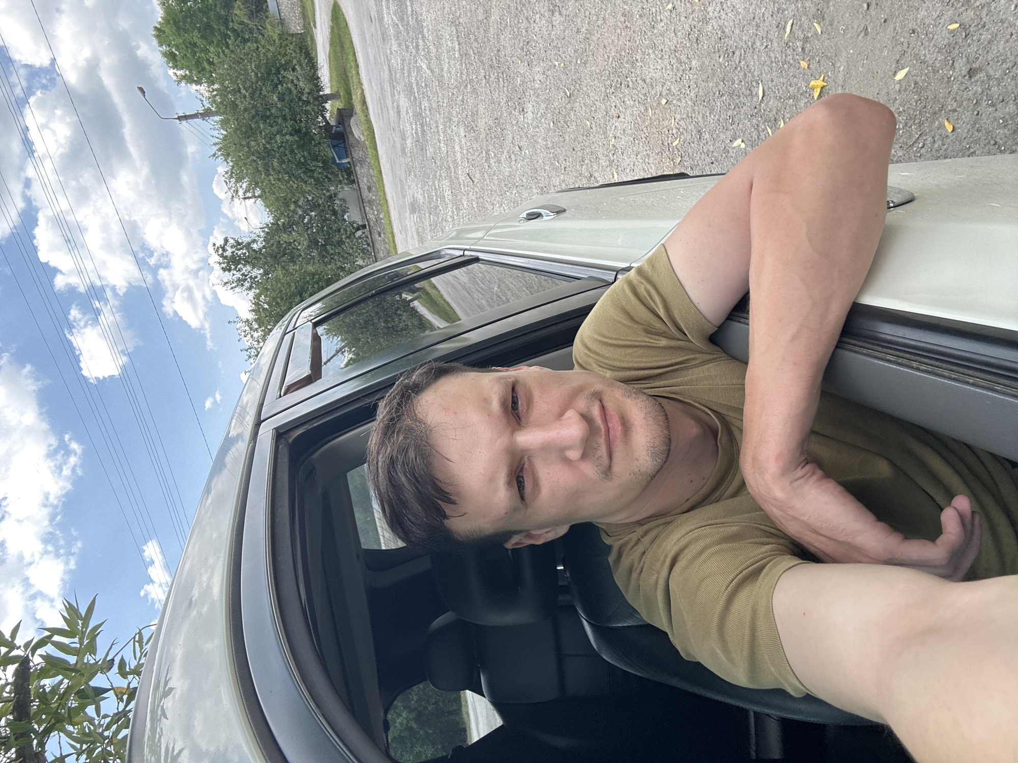 A man is sitting in the driver's seat of a car, with his arm resting on the open car door. He is looking at the camera. The car is parked on a gravel lot next to a paved road. In the background, there are trees, a street lamp, and a blue tarp. The sky is cloudy. The image was taken in Makiv, Ukraine.