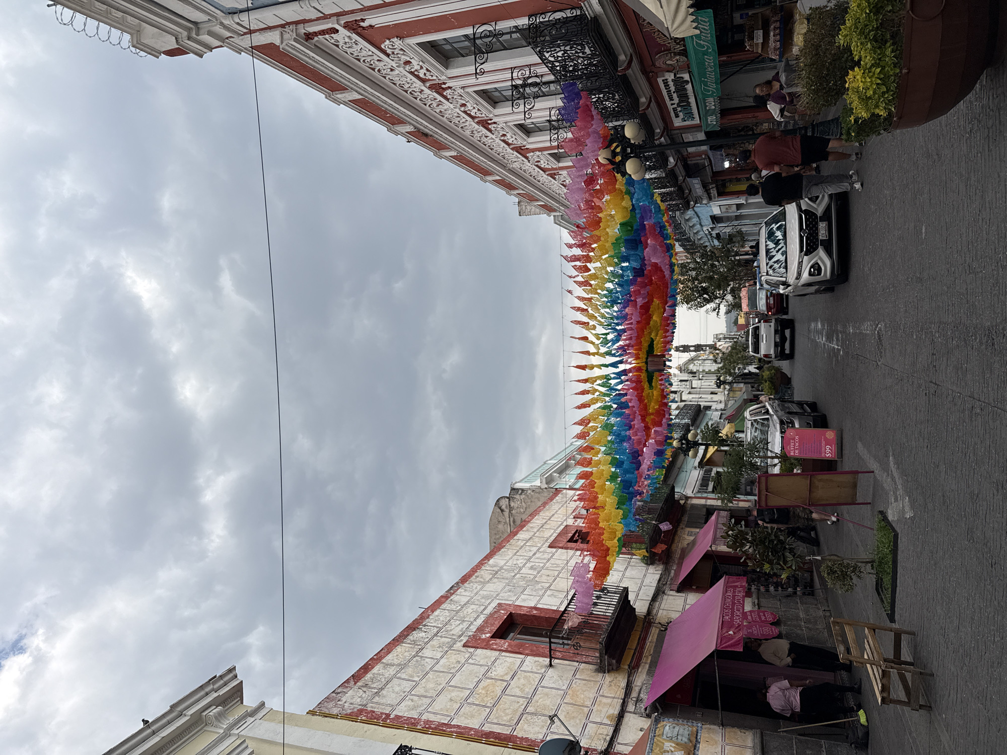This full-shot image presents a bustling street scene within a historic urban setting, characterized by traditional architecture and vibrant cultural decorations. The perspective is slightly angled, looking down a street lined with multi-story buildings under an overcast sky.
Dominating the overhead space is a long garland of colorful papel picado, traditional Mexican perforated paper flags, stretched across the street. These flags, arranged in a rainbow spectrum, create a festive canopy.
The buildings on either side exhibit ornate facades with intricate details, balconies, and decorative cornices. On the left, a building features red trim and white patterns, with an aged, textured wall displaying a tiled design at its lower level. On the right, storefronts with awnings line the sidewalk, housing various businesses.
Several people are visible walking and standing on the sidewalks. A white SUV with the license plate "EK-332-6" is parked on the street, and other vehicles can be seen further down the road. Potted plants and streetlights contribute to the urban environment.
Visible text on signs includes "TACOS CHINGONES" and "SABORCITO CORAZÓN," advertising a taco shop. Another sign promotes a "BUFFET DE TACOS" for "$99," likely in Mexican pesos. Other partial texts like "Santo Dom" and "STO. DON Talavera Frida" hint at local businesses or landmarks. The cloudy weather suggests a daytime scene, while the decorations evoke a lively, cultural atmosphere characteristic of places like Puebla, Mexico.