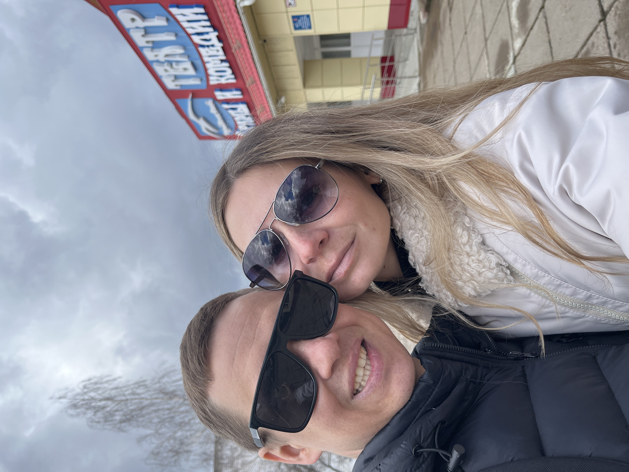 The image shows a selfie of a man and a woman taken outside a building in Chaikovsky, Russia. The woman has long blonde hair and is wearing aviator sunglasses and a white jacket. The man has short hair and is wearing black sunglasses and a black puffer jacket. They are both smiling. The background features a building with a red and blue sign with Cyrillic letters, which reads "ТЕАТР И КОМПАНИЯ" (Theatre and Company) and a second building with a yellowish facade and a gray sky above. The ground appears to be paved with concrete or stone.