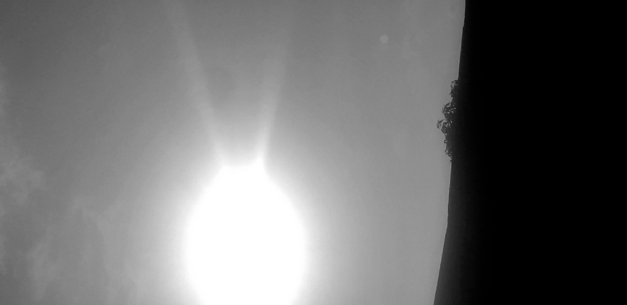The image shows an outdoor scene in Narayi, Nigeria. The sun is brightly visible through a hazy sky, creating a strong glare. A dark, undefined object, potentially a building or tree, occupies the right side of the frame. Some foliage can be distinguished on the edge of this dark object. The rest of the image is filled with sky and clouds, with some variations in gray tones indicating different levels of light and cloud density.