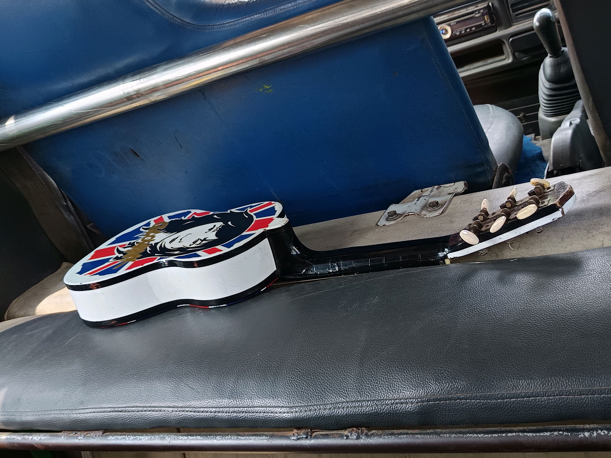 In Karangnangka, Indonesia, a white guitar with a Union Jack pattern and a lion's head emblem is placed on a dark gray, textured seat, possibly in a vehicle. The guitar's neck extends to the right, showing tuning pegs and a fretboard. The background includes a blue seat, a metal bar, and parts of a vehicle's interior, such as a gear shift and a radio. The scene is illuminated by natural light, casting shadows and highlighting the textures of the materials.