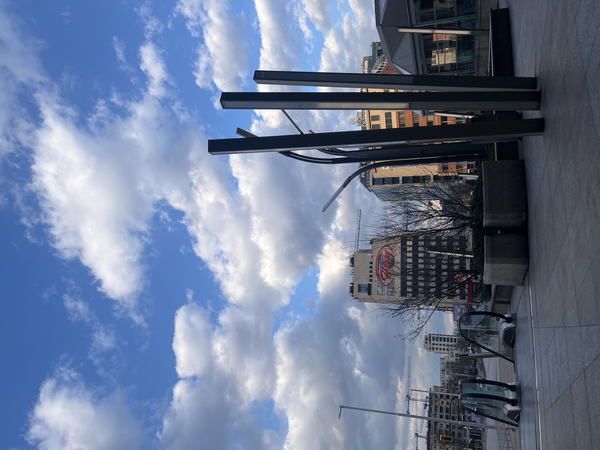 The image depicts an outdoor scene in Sofia, Bulgaria. The view is directed towards a cityscape featuring a mix of modern and older buildings. In the foreground, there are architectural elements, including dark, rectangular pillars and metallic curved structures, likely part of a building or public space design. The ground is paved with grey tiles.
Further into the view, there is a building with a prominent "Dessy" sign in red. Cranes are visible in the distance, indicating ongoing construction in the area. The sky is blue with puffy white clouds, suggesting a sunny day. A bare tree is also visible. An escalator entrance is visible near the bottom of the image.