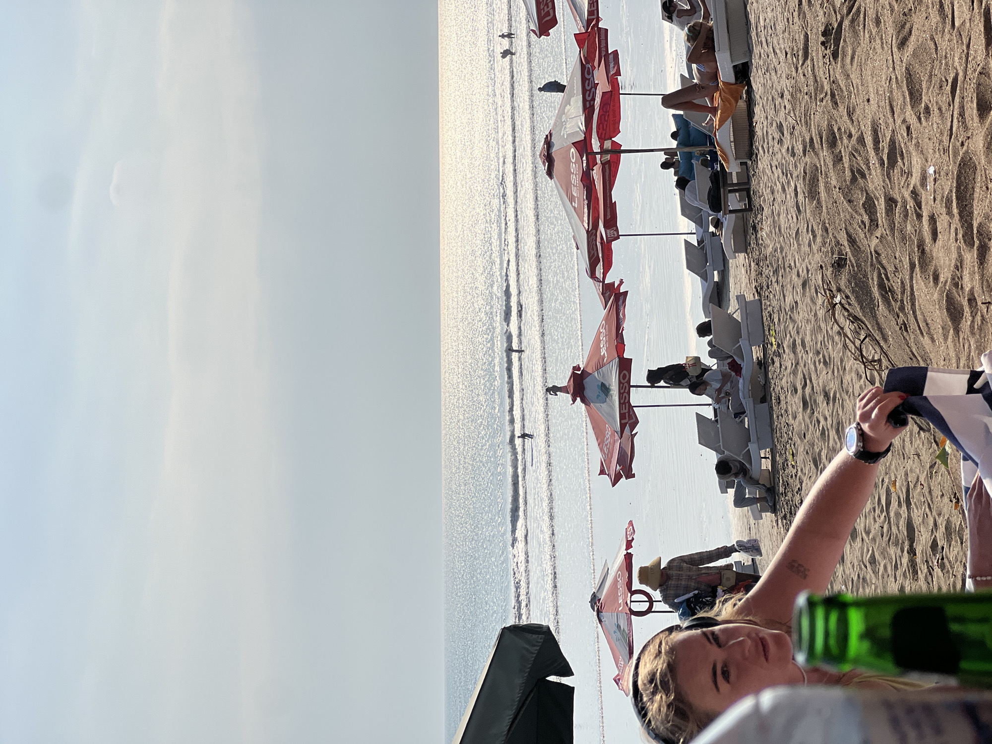 The image depicts a bustling beach scene in Kuta, Indonesia. In the foreground, a woman with blonde hair and headphones is partially visible, looking to her left. Her right arm is extended, and she is holding a dark and white striped cloth. A green glass bottle is positioned in front of her, slightly out of focus. Behind her, beach chairs are set up on the sandy shore, some occupied by people lounging. Several red and white umbrellas, branded with "LESSO," provide shade over the chairs.
In the middle ground, the ocean stretches out, with gentle waves breaking on the shore. The water is sparkling under the sunlight. A few individuals are in the water, with one person seen surfing or paddleboarding in the distance. Other people are visible walking or standing near the water's edge.
The background consists of the sea and a hazy sky with scattered clouds, suggesting it might be late afternoon. The overall impression is a lively beach environment characteristic of Kuta, a popular tourist destination in Bali, Indonesia, known for its beaches and surf.