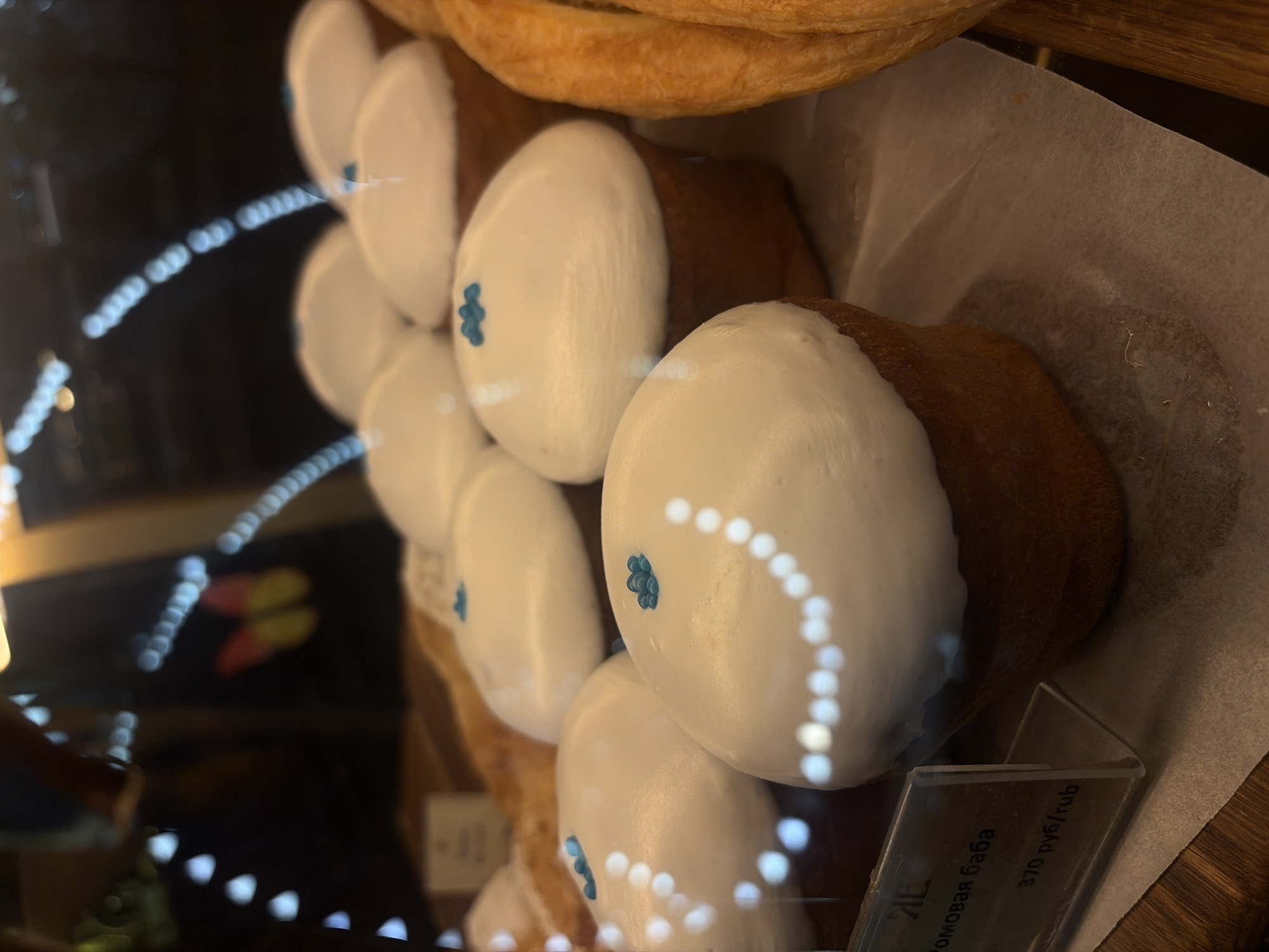 The image shows a close-up of several pastries in what appears to be a bakery display case. The pastries are individual servings, topped with white frosting and decorated with a few small blue sprinkles. Some of the frosting has reflections from lights above, creating a bokeh effect. In the foreground, to the left, is a price tag made of clear plastic. Text on the tag, in Russian, reads "РОМОВАЯ БАБА" and "370 руб/rub". The overall lighting is dim, with artificial lights highlighting the pastries. In the background, a blurred image of a yellow and pink butterfly decoration can be seen. The pastry is a "Romovaya Baba" which translates to "Rum Baba" or "Baba au Rhum", a traditional yeast cake soaked in rum syrup. Given the Russian text and the type of pastry, this scene is likely from a bakery in Saint Petersburg, Russia.