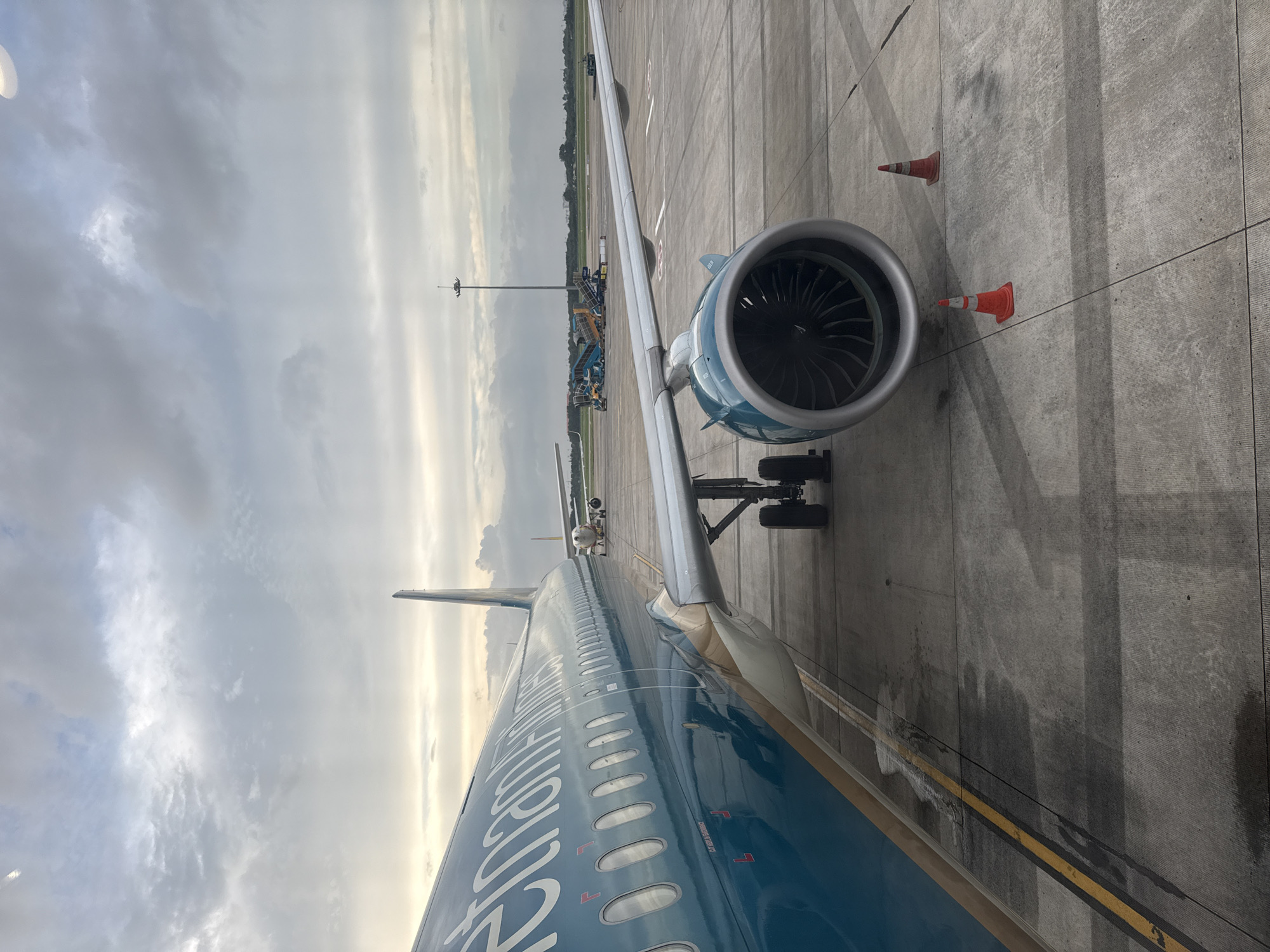 The image shows the side of a Vietnam Airlines airplane parked on an airport tarmac. The airplane is a large, commercial jet with a teal fuselage and gold accents. Its engines are visible, along with the landing gear. In the background, other airplanes, airport service vehicles, and a tall light tower are present. The scene is set at an airport in Ho Chi Minh City, Vietnam, under a cloudy sky. Two orange traffic cones are placed on the tarmac near the engine.