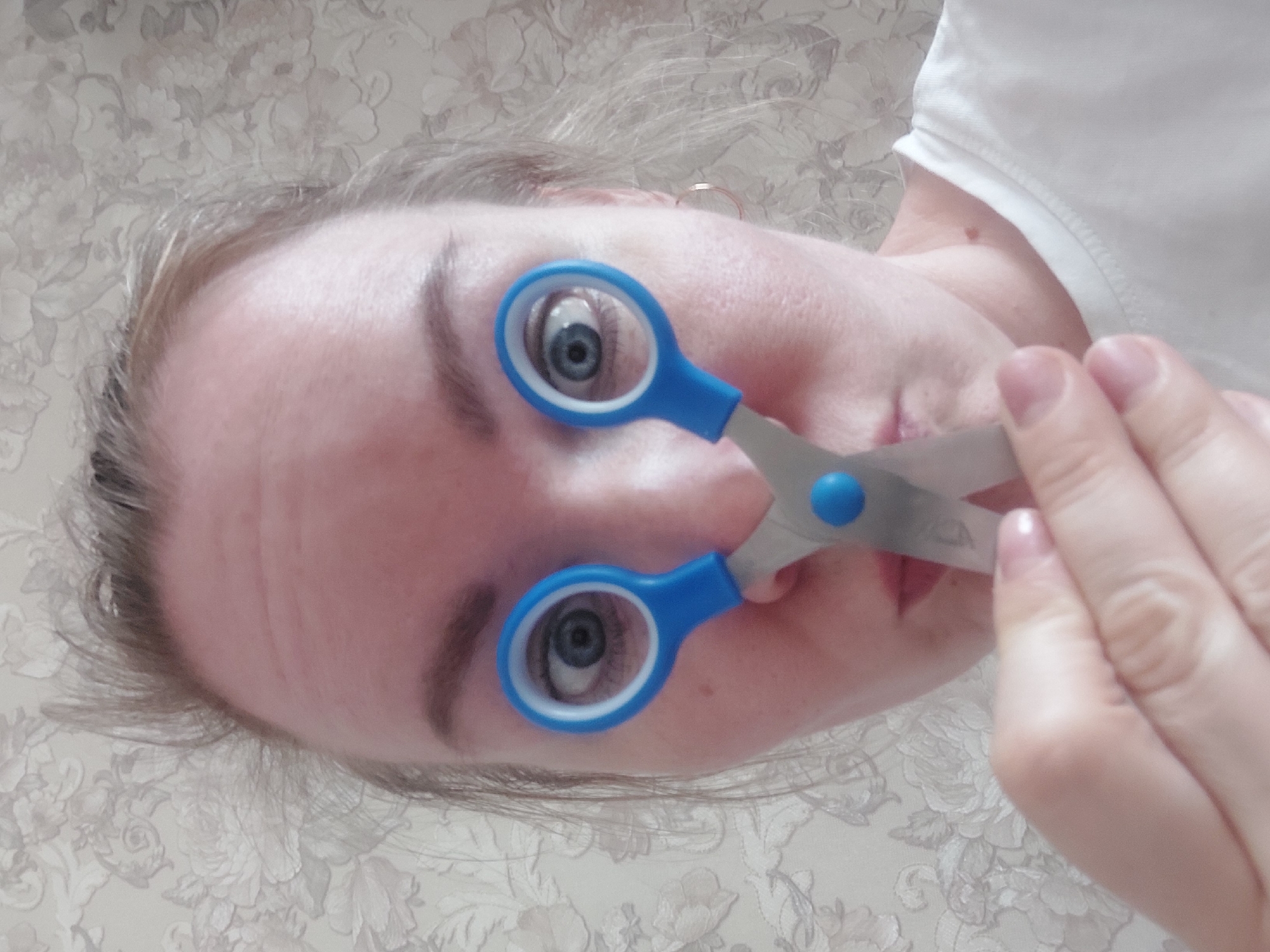 A woman's face is positioned diagonally against a floral wallpaper background. She holds a pair of blue-handled scissors with the eye-holes framing her eyes. Her right hand steadies the scissors. The scene is most likely indoors. The location of Chaikovsky, Russia, is not verifiable from the image.