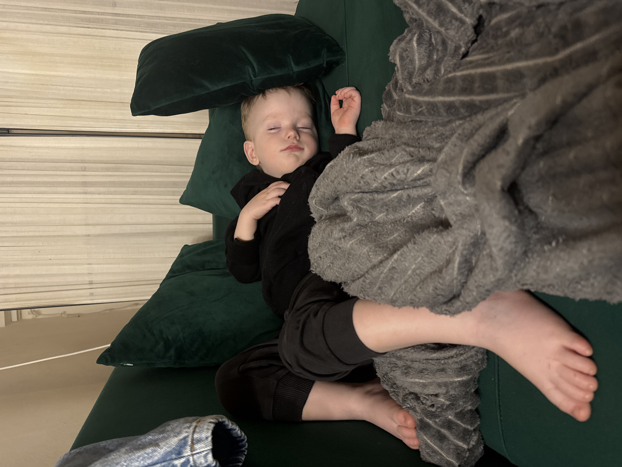 A young child is asleep on a dark green sofa. The child is lying on their back with closed eyes and a hand resting on their chest. The child is wearing a black hooded sweatshirt and dark pants. A large gray blanket partially covers the child's legs. The child's feet are visible outside the blanket. Dark green pillows are positioned behind the child's head. A blue denim object is in the lower left of the frame. The background consists of a wall with horizontal panels. It is impossible to determine if the image was taken in the city of Saint Petersburg, Russia.