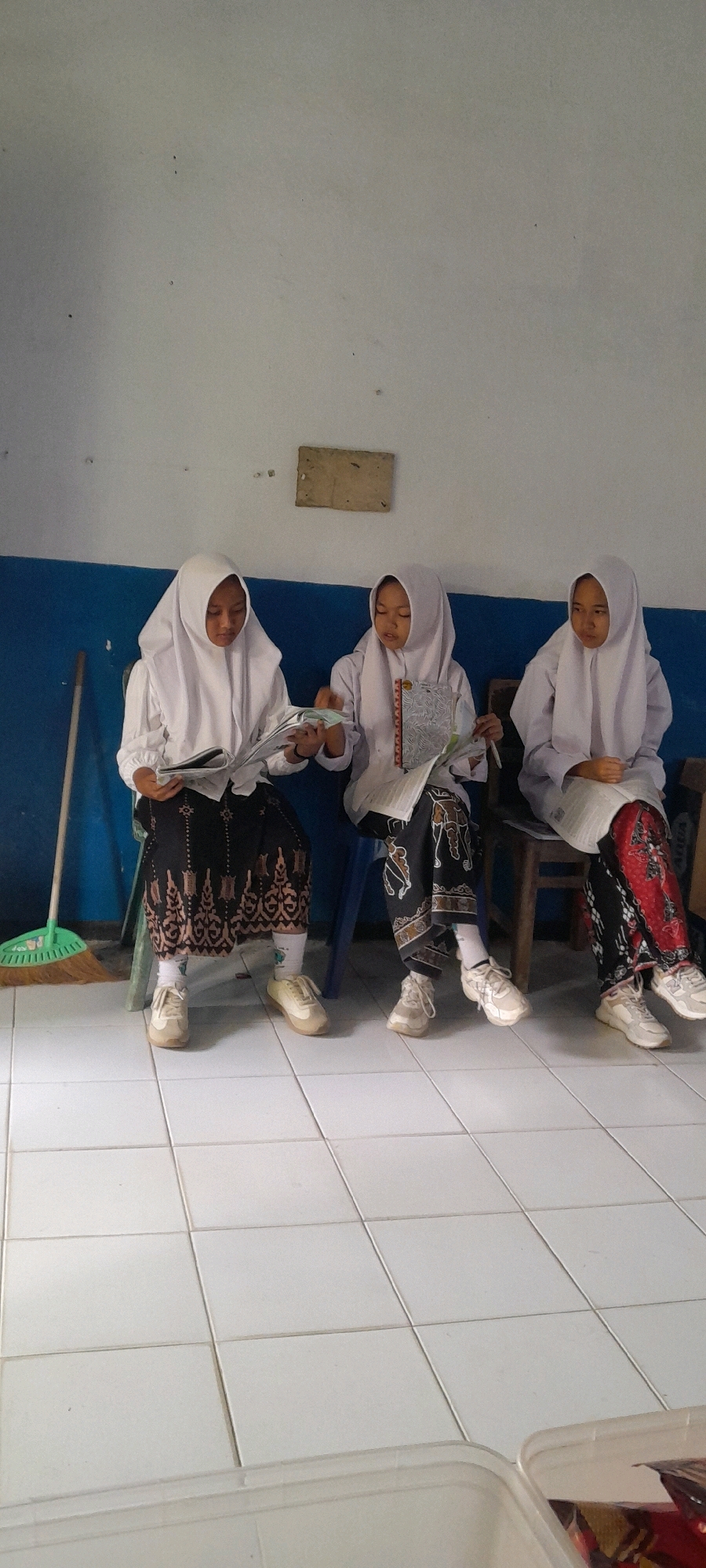 Three young girls wearing white hijabs and long-sleeved white shirts are seated on chairs in front of a blue and white wall. They are all reading from books or magazines. The girl on the left is wearing a black skirt with a patterned hem, white socks, and light-colored sneakers. She is holding a large open book or magazine. The middle girl is wearing a patterned skirt, white socks, and light-colored sneakers, and she is holding an open book with a colorful cover. The girl on the right is also wearing a patterned skirt, white socks, and light-colored sneakers, and she is holding an open book. A broom with a green handle is visible on the left side of the image. The floor is tiled with white square tiles. A rectangular piece of material is attached to the wall above the girls. This scene appears to be indoors, possibly a classroom or community space, in Tahunan, Indonesia, as suggested by the context.