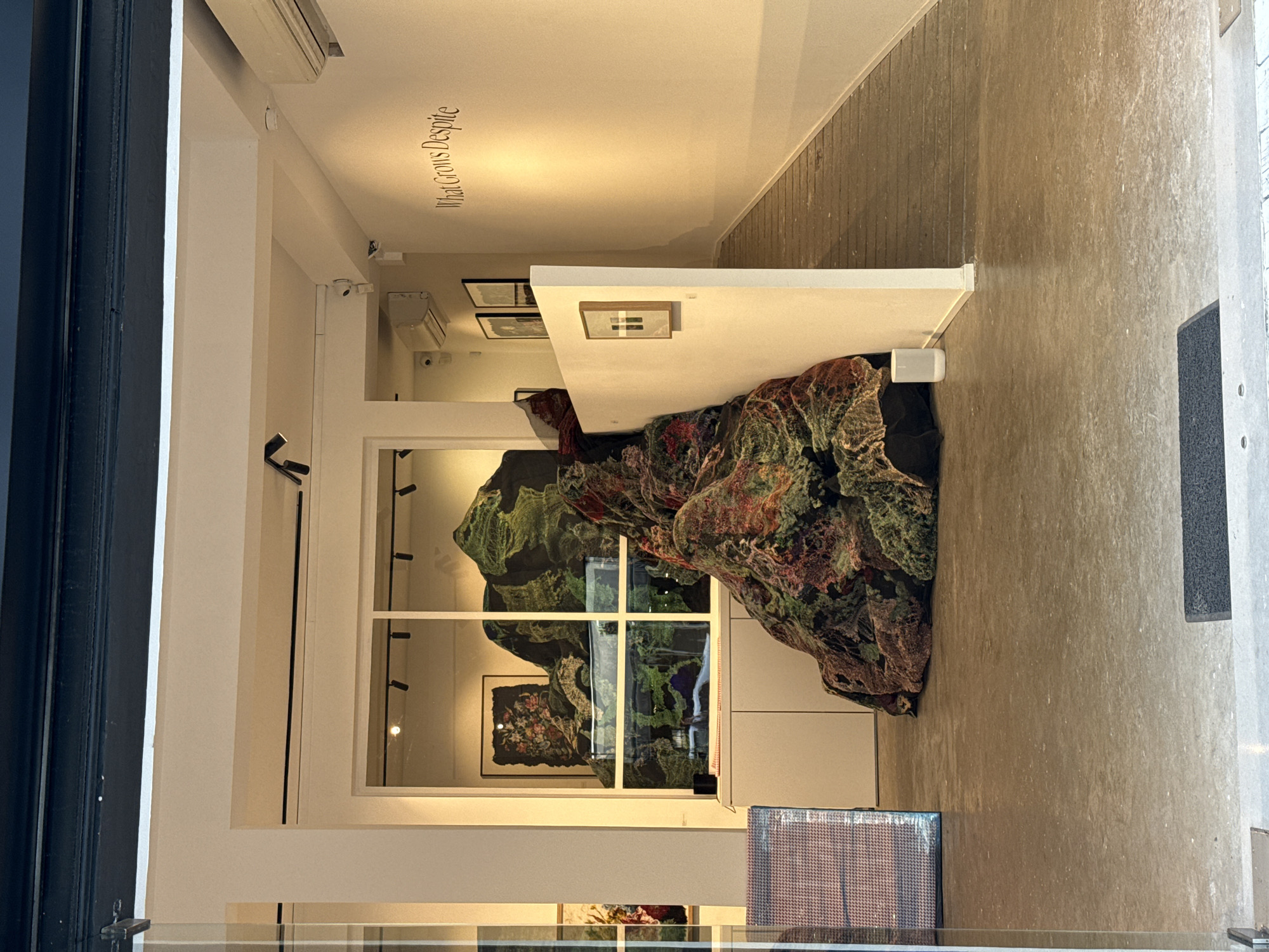 The photograph shows an art installation in a gallery located in Tel-Aviv, Israel. The installation consists of a large, sculpted form with a camouflage-like pattern of green, red, and black shades. It spills over from a white platform and occupies a significant portion of the gallery space. In the background, through a window or a display case, is a framed artwork showing floral patterns. The gallery's walls are white, and there is text "What Grows Despite" visible on the upper portion of one wall. The floor is a gray concrete surface. The entrance or exit of the gallery is partially visible on the sides of the frame.