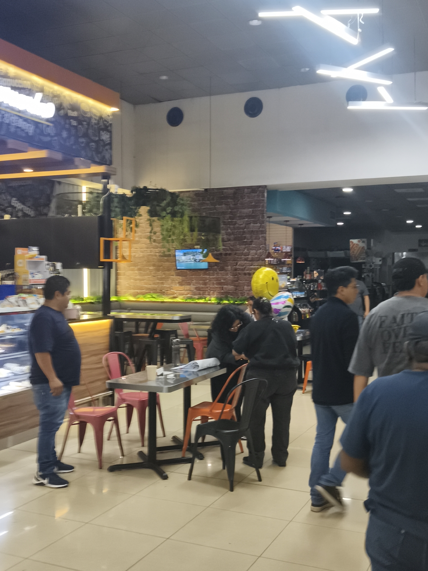 The image shows the interior of a restaurant in Guatemala City, Guatemala. The flooring is a light-colored tile, and the lighting comes from ceiling-mounted fluorescent fixtures. There are several people visible, including customers and staff. To the left is a display case with food items. Behind this is a man in a blue t-shirt and jeans.
In the middle of the image, two individuals appear to be interacting at a table; both are wearing darker clothing. A yellow smiley-face balloon and a few other balloons are visible in the background behind the people at the table. To the right of this group, two men are walking. The man closest to the camera is wearing a black shirt and jeans, while the man behind him is wearing a grey t-shirt. Various tables and chairs are scattered throughout the space. A brick-like wall with greenery on top and a television screen are visible in the background.