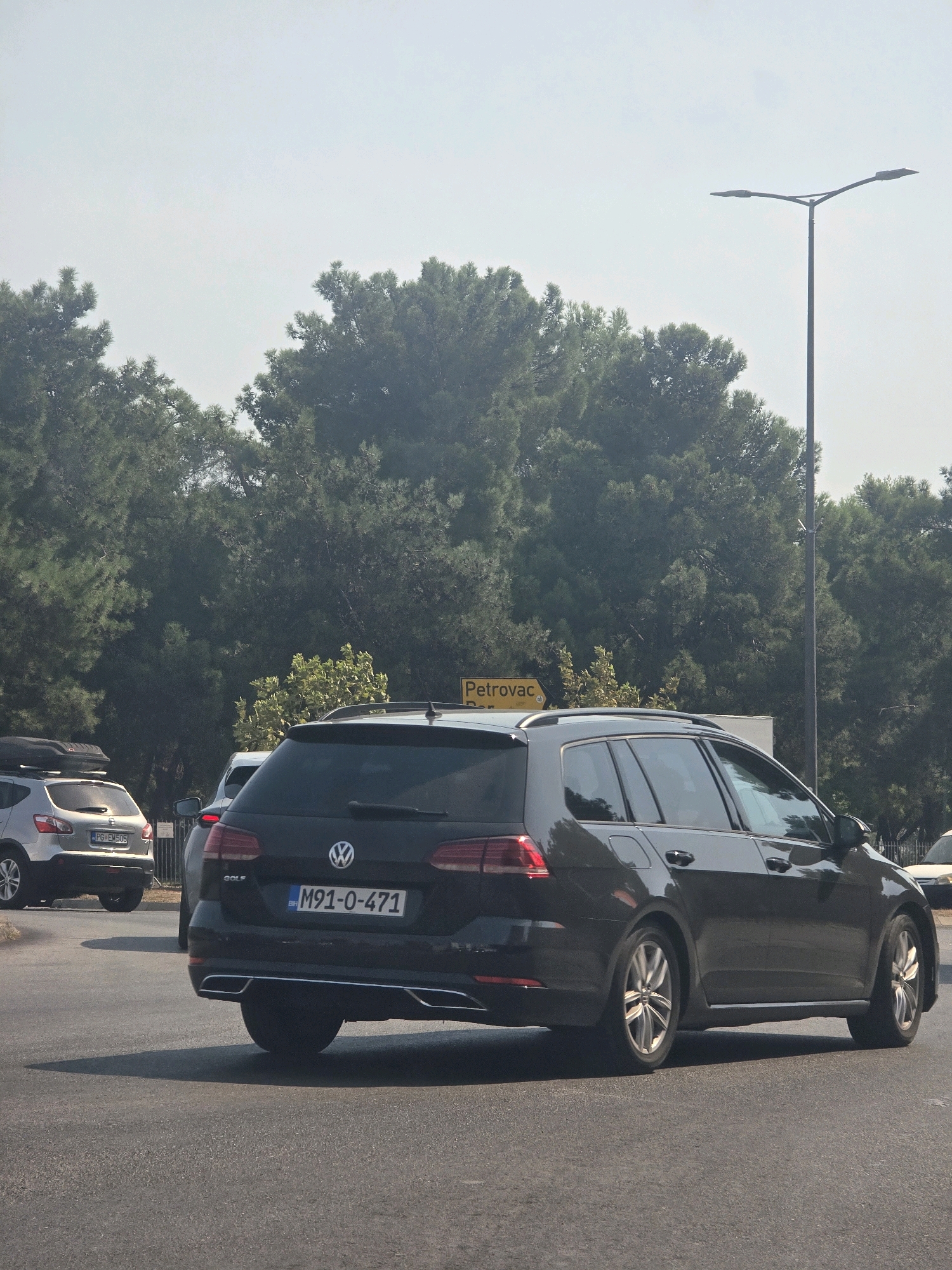 In Podgorica, Montenegro, on a sunny day, a dark Volkswagen Golf station wagon is seen from the rear passenger side, driving on asphalt. The car's license plate reads "M91-0-471". To the left, a silver Nissan Qashqai with a roof box and license plate "PG-EM505" is visible, also in traffic. Behind the cars, a dense line of green coniferous trees creates a backdrop, with a bright sky overhead. A tall street lamp stands to the right of the frame. A yellow road sign indicating "Petrovac" is partially visible amongst the trees.