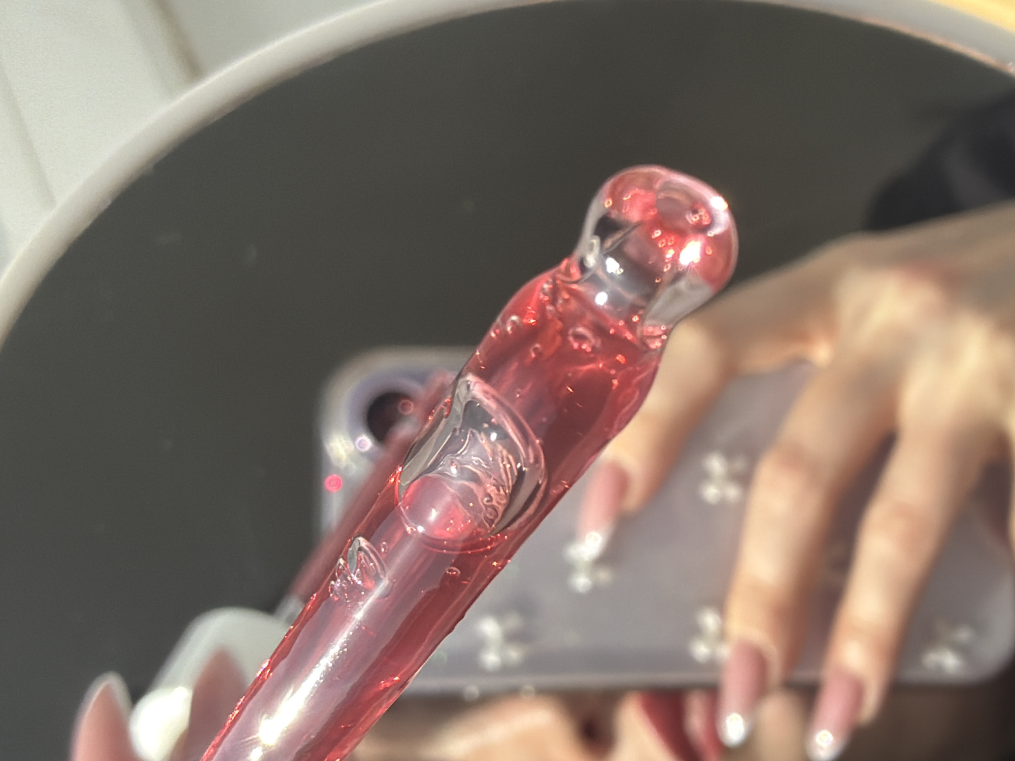 The image captures a close-up, mirrored reflection of a scene in Saint Petersburg, Russia. A person's hand, featuring long, oval-shaped nails with a pink base and white French tips, holds a transparent glass dropper horizontally. The dropper is filled with a translucent, shimmering red liquid containing numerous small bubbles. Partially visible in the lower-left reflection are the person's lips, adorned with dark red lipstick, and a portion of their nose. The individual's other hand holds a smartphone with a light-colored, possibly translucent, case embellished with multiple small, shiny, bow-shaped or star-shaped decorative elements. A segment of the phone's camera module is also visible in the reflection. The environment is an indoor setting, characterized by a dark, indistinct background reflected within the mirror, which itself has a discernible white, curved edge on the right.