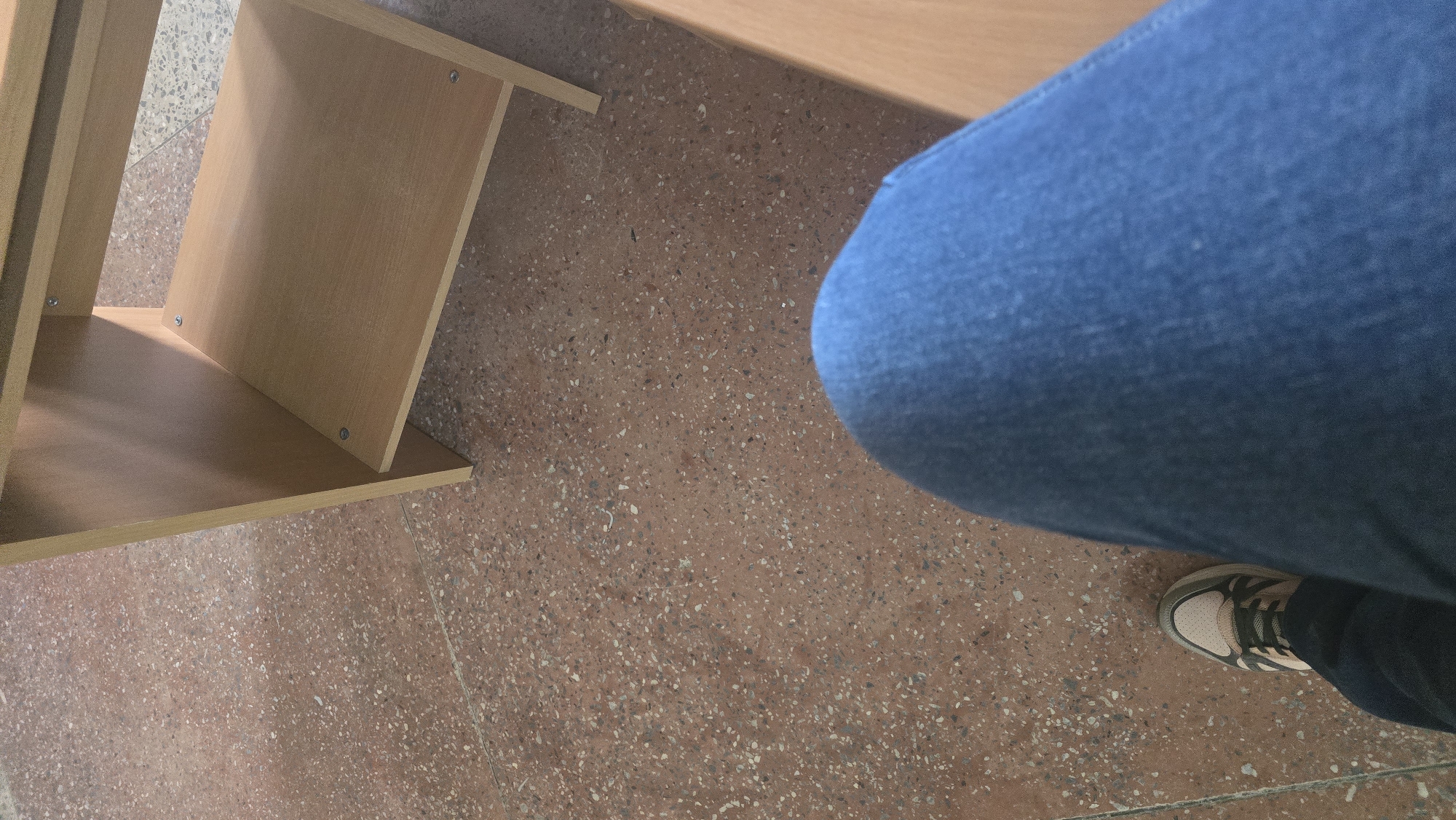 The image shows a person's legs, clad in blue denim jeans and a sneaker, resting on a speckled brown floor. To the right, a light brown wooden shelf unit is visible, with two shelves. The scene appears to be indoors, possibly in a classroom or office setting. The lighting suggests it could be daytime. No specific landmarks or recognizable features place the image definitively in Ukhta, Russia.