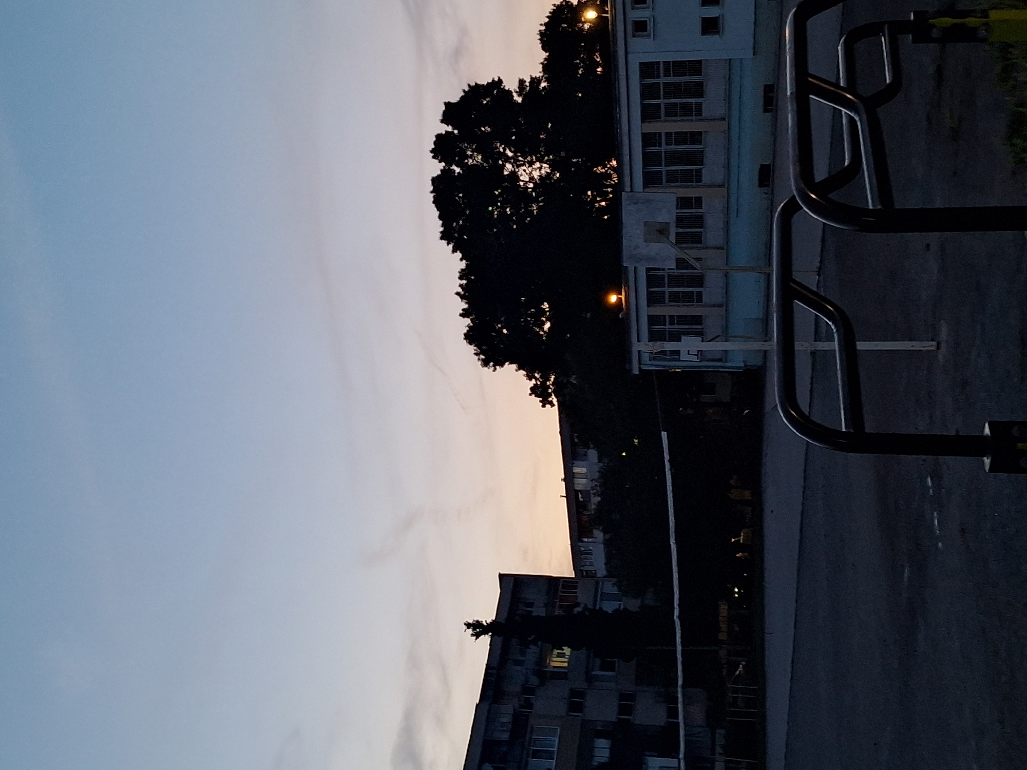The image captures an outdoor scene at dusk in Varna, Bulgaria. In the foreground, dark metal railings of playground equipment are visible. Beyond the railings, a paved area, likely a sports court, stretches into the mid-ground. A dark net, possibly for volleyball, is strung across the court. In the background, silhouetted against the pale twilight sky, are several buildings and trees. On the left, a multi-story apartment building with visible windows stands prominently. To the right of the apartment building and central in the frame, a large, dense tree with intricate branches is silhouetted. Further to the right, a low-rise building with multiple windows and a basketball hoop mounted on its exterior is visible. Two bright yellow lights are illuminated on the roofline of this building. The sky is a gradient of pale blue at the top, transitioning to a soft orange and pink hue near the horizon, indicating the end of daylight. The overall atmosphere is calm and serene, characteristic of early evening in an urban setting.
